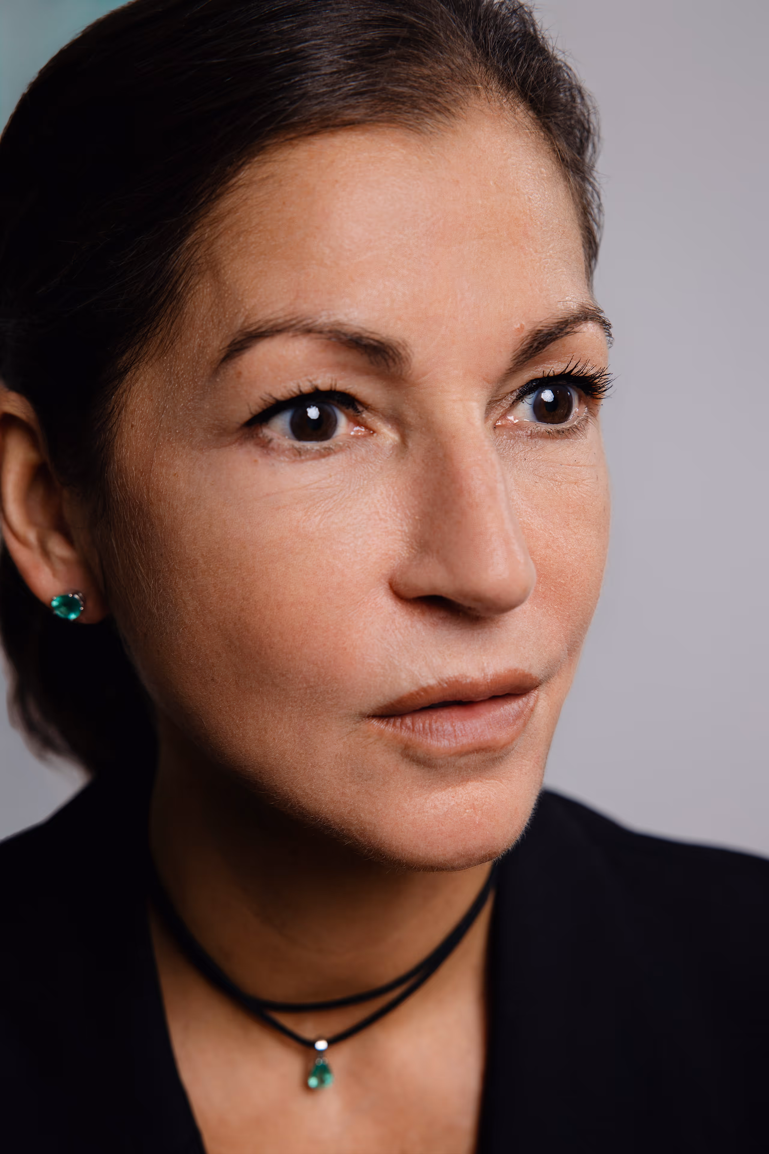 Porträt einer Frau mit dunkelhaarigem zurückgebundenem Haar, die grüne Ohrringe und eine passende Halskette trägt.