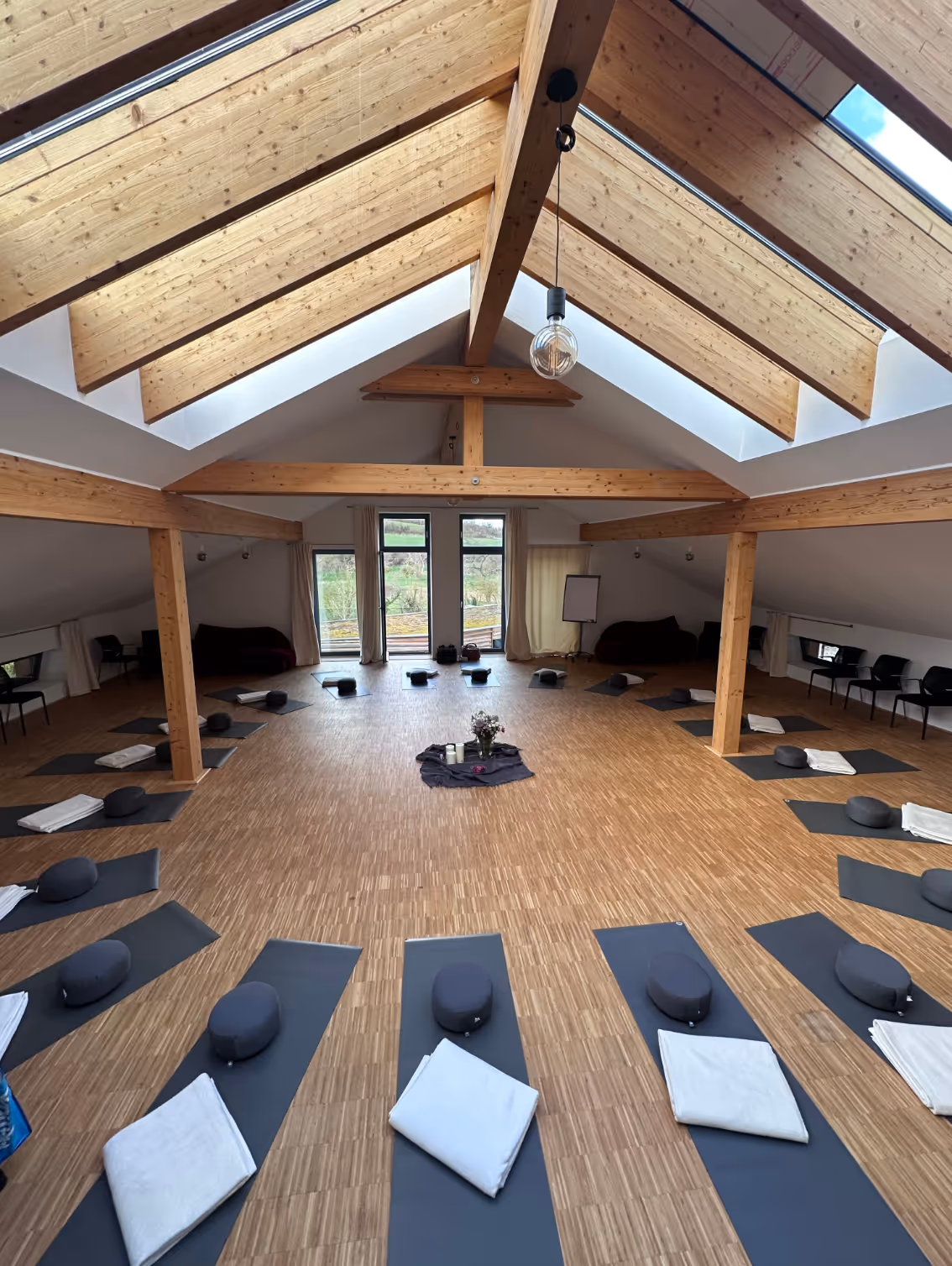 Ein heller und geräumiger Meditationsraum mit Holzbalken und großen Fenstern, ausgestattete Yogamatten, Meditationskissen und Decken im Kreis angeordnet.