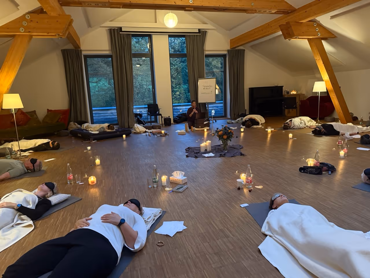 Menschen liegen mit Augenbinden auf Yogamatten in einem ruhigen, kerzenbeleuchteten Raum mit Holzbalken und großen Fenstern.