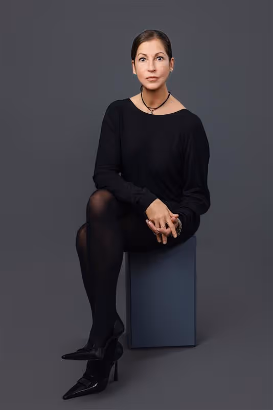 Porträt einer Frau mit zurückgebundenen braunen Haaren, die in schwarzer Kleidung sitzt vor grauem Hintergrund.