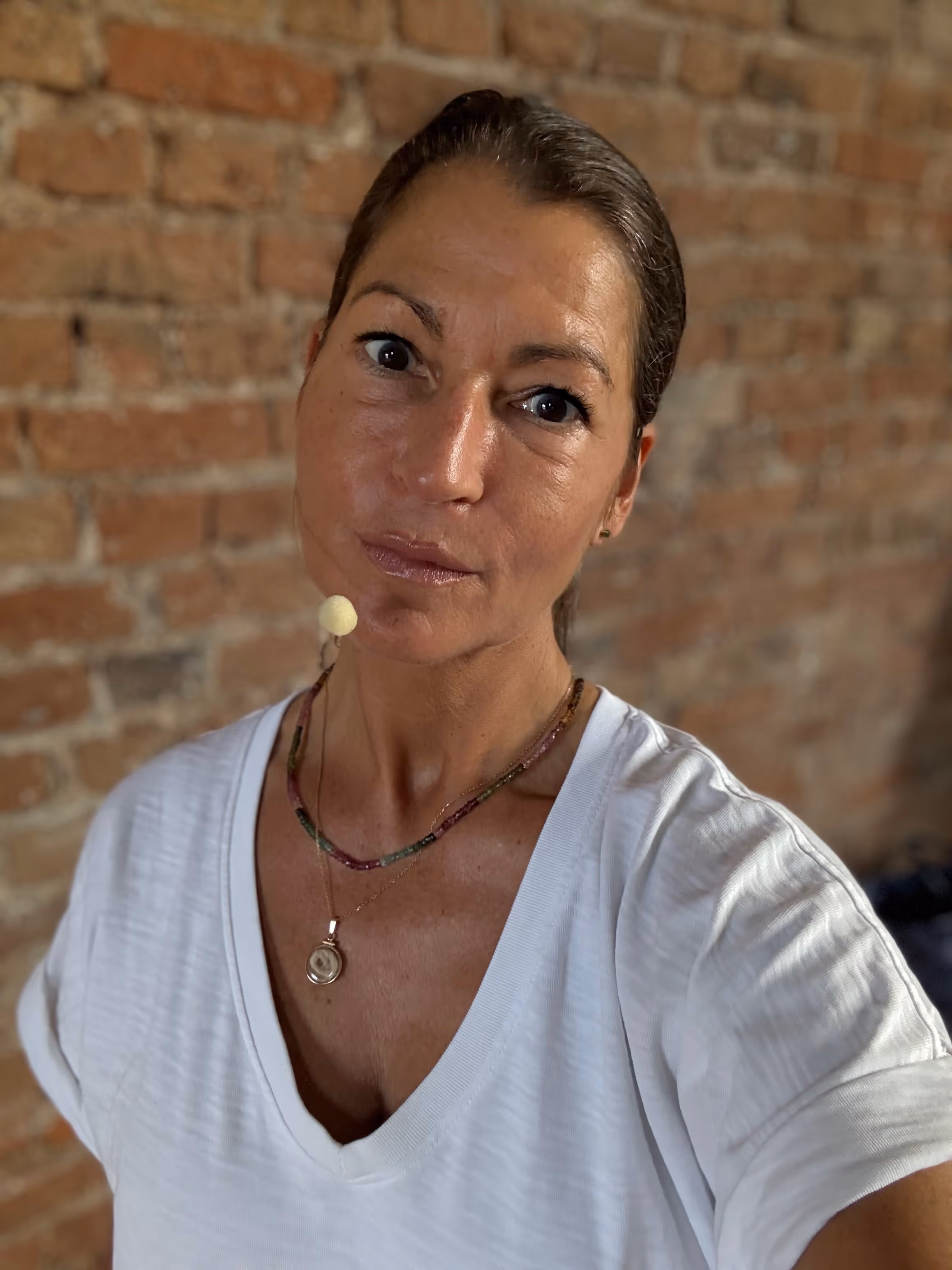 Frau mit braunem zurückgebundenem Haar trägt weißes T-Shirt und zwei Halsketten, stehend vor einer Ziegelsteinwand.