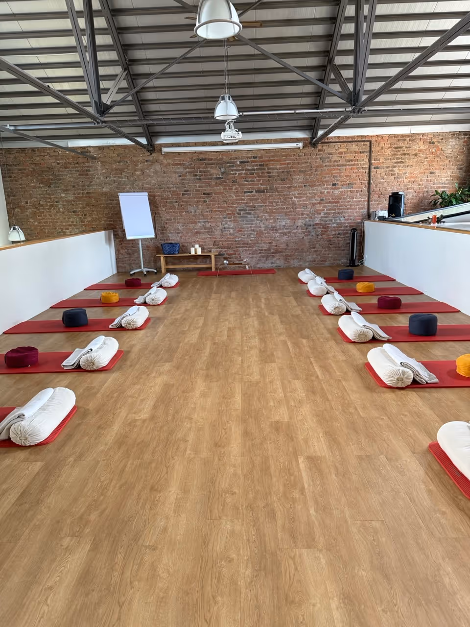 Yoga- oder Meditationsraum mit Holzfußboden, roten Matten, weißen Kissen und farbigen Meditationspolstern entlang der Wände, Backsteinwand und Deckenlampen.