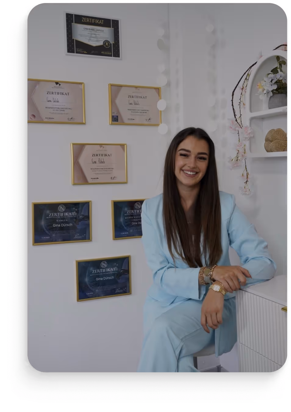Junge Frau in hellblauem Anzug sitzt lächelnd vor einer Wand mit mehreren zertifizierten Urkunden.