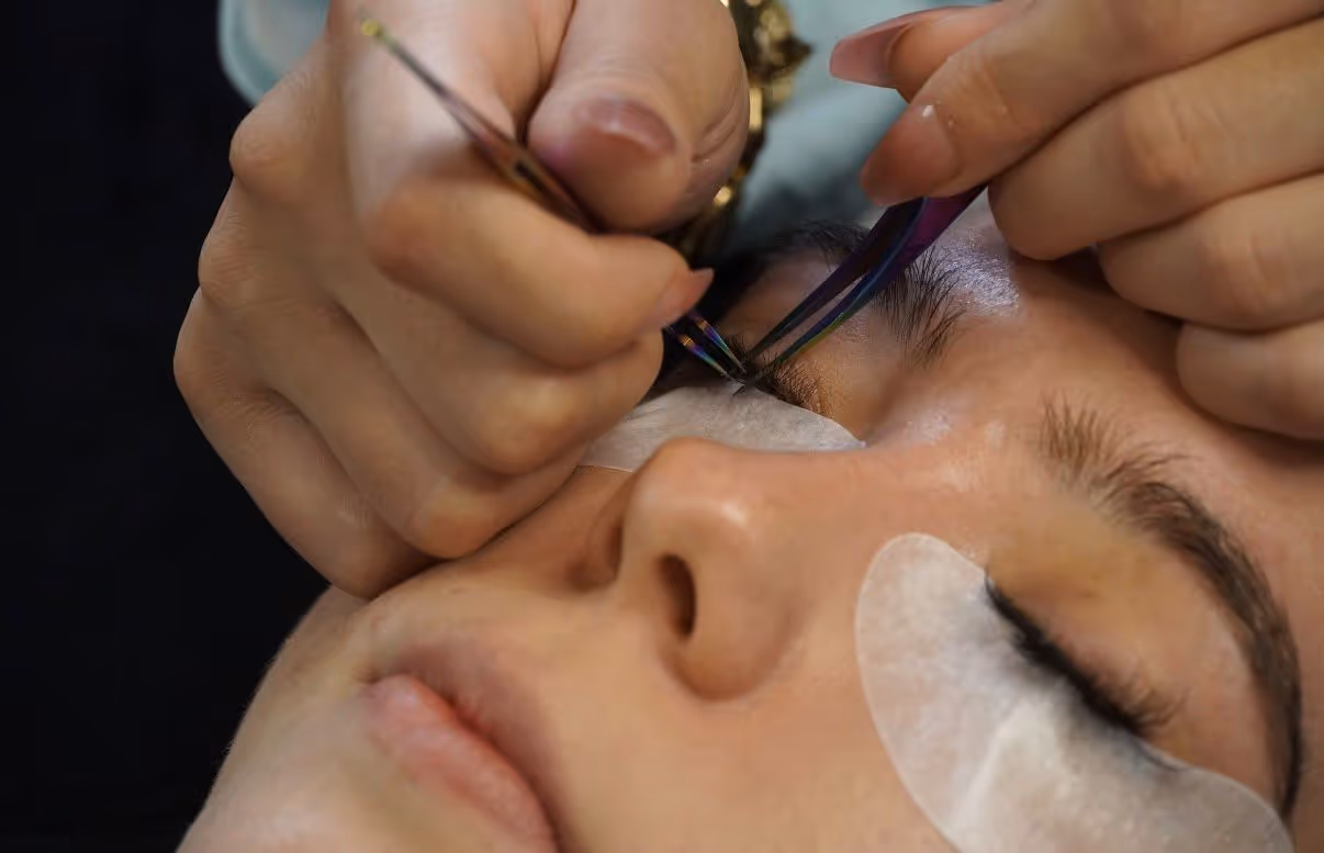 Kosmetikerin trägt eine Behandlung mit einem Pinsel auf die geschlossenen Wimpern einer liegenden Frau auf, die unter den Augen Pads trägt.