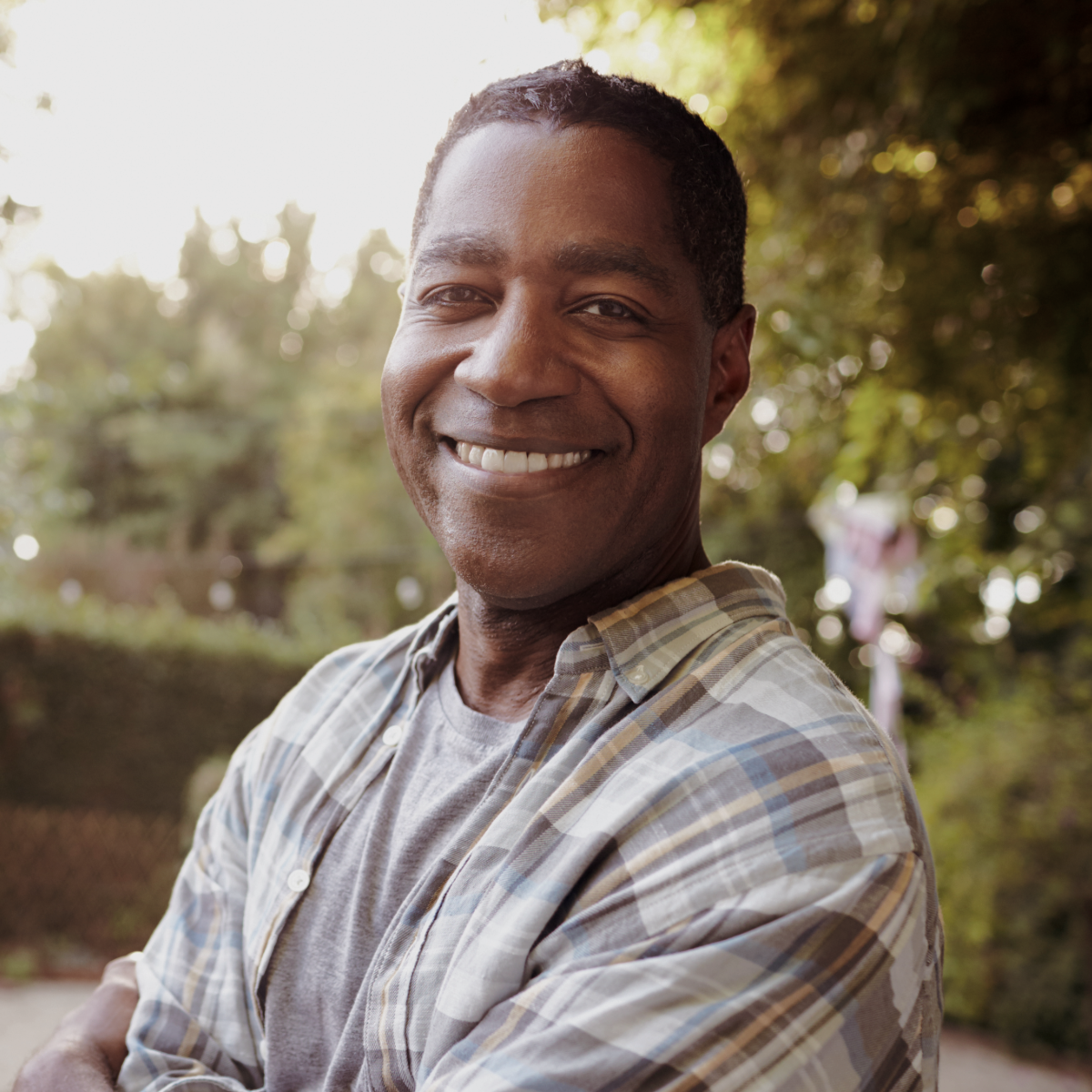 Smiling man wearing a plaid shirt stands outdoors with arms crossed in a sunlit garden.