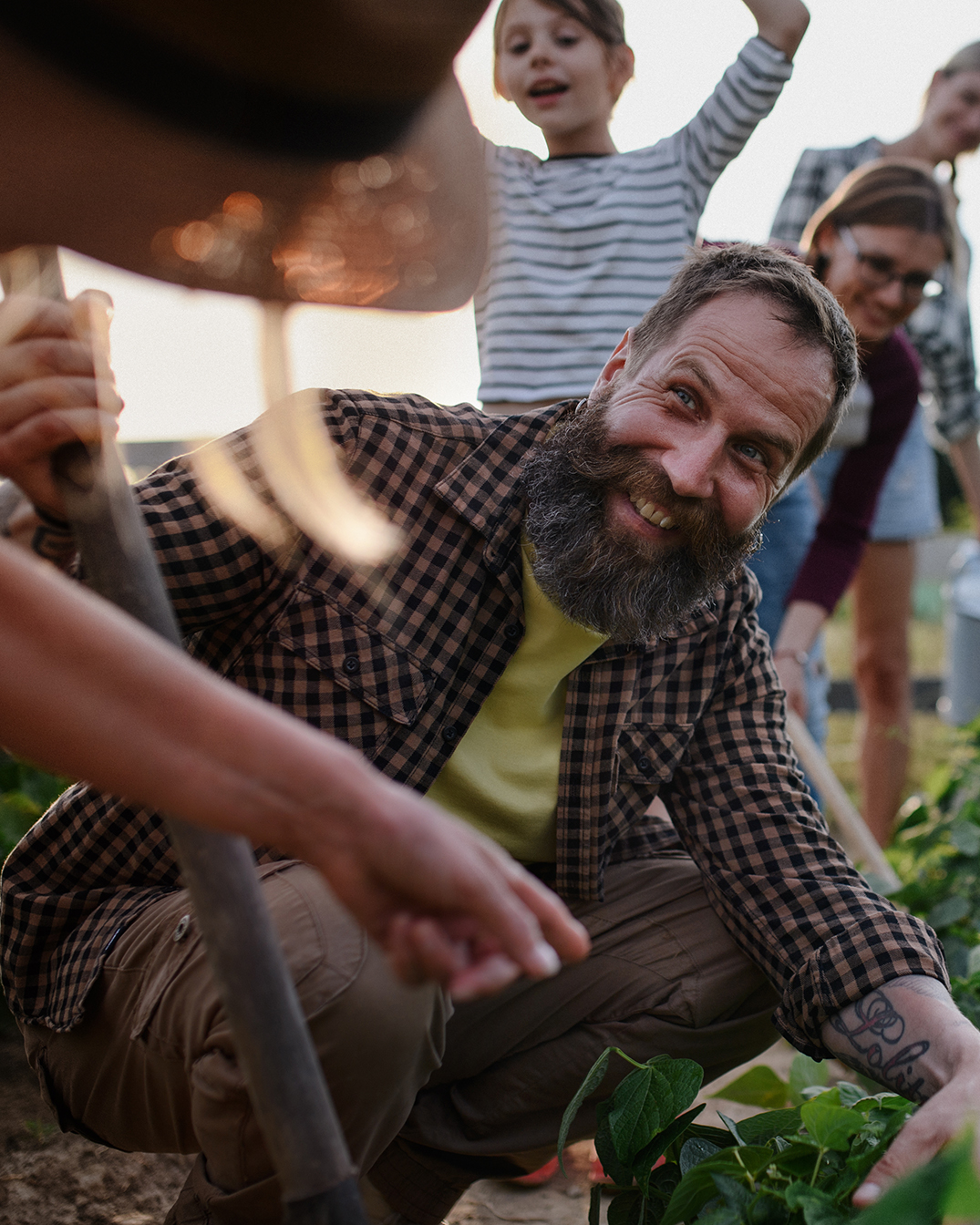 A bearded man in a checkered shirt smiles while gardening outdoors, with a child and two adults in the background.