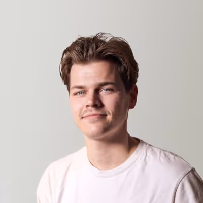 Young man with short brown hair and a slight mustache, wearing a white t-shirt, smiling against a light gray background.