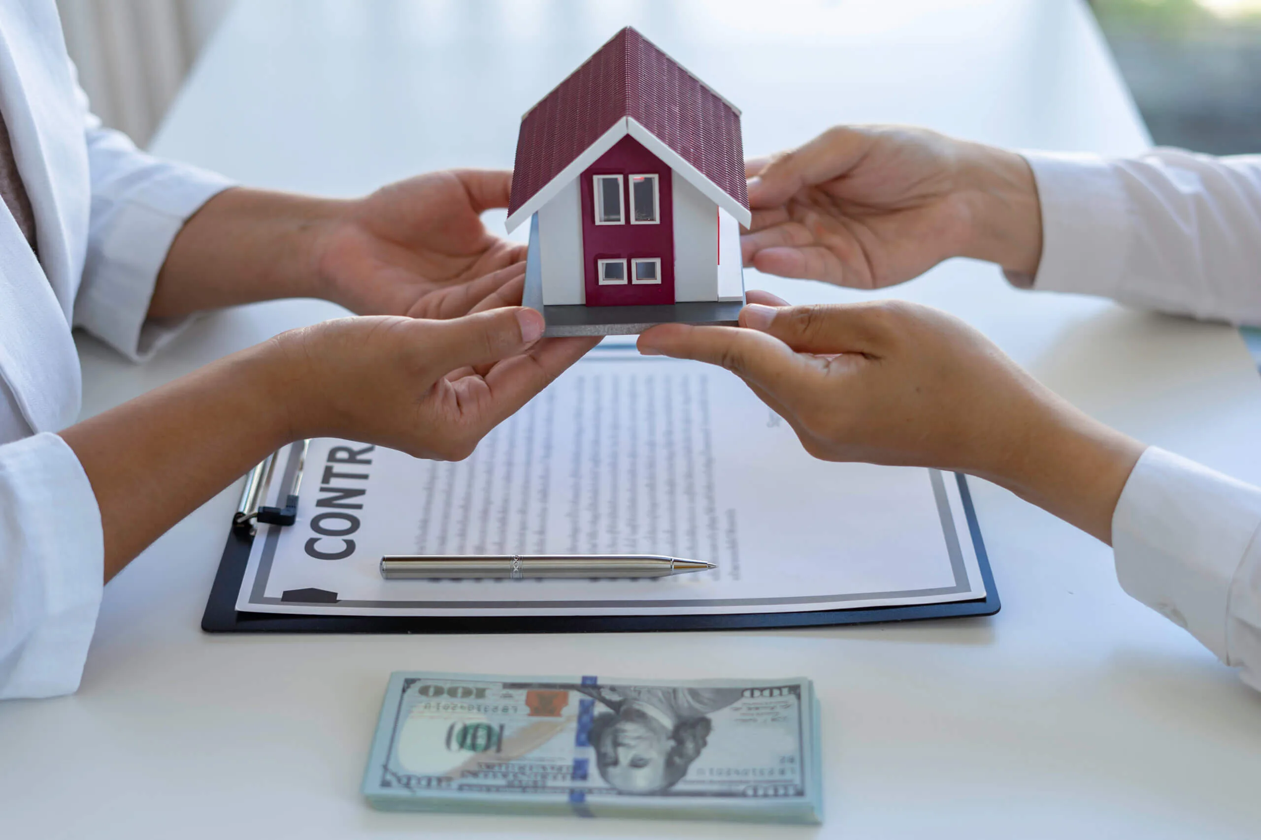 Two hands holding small house, money and contract on table