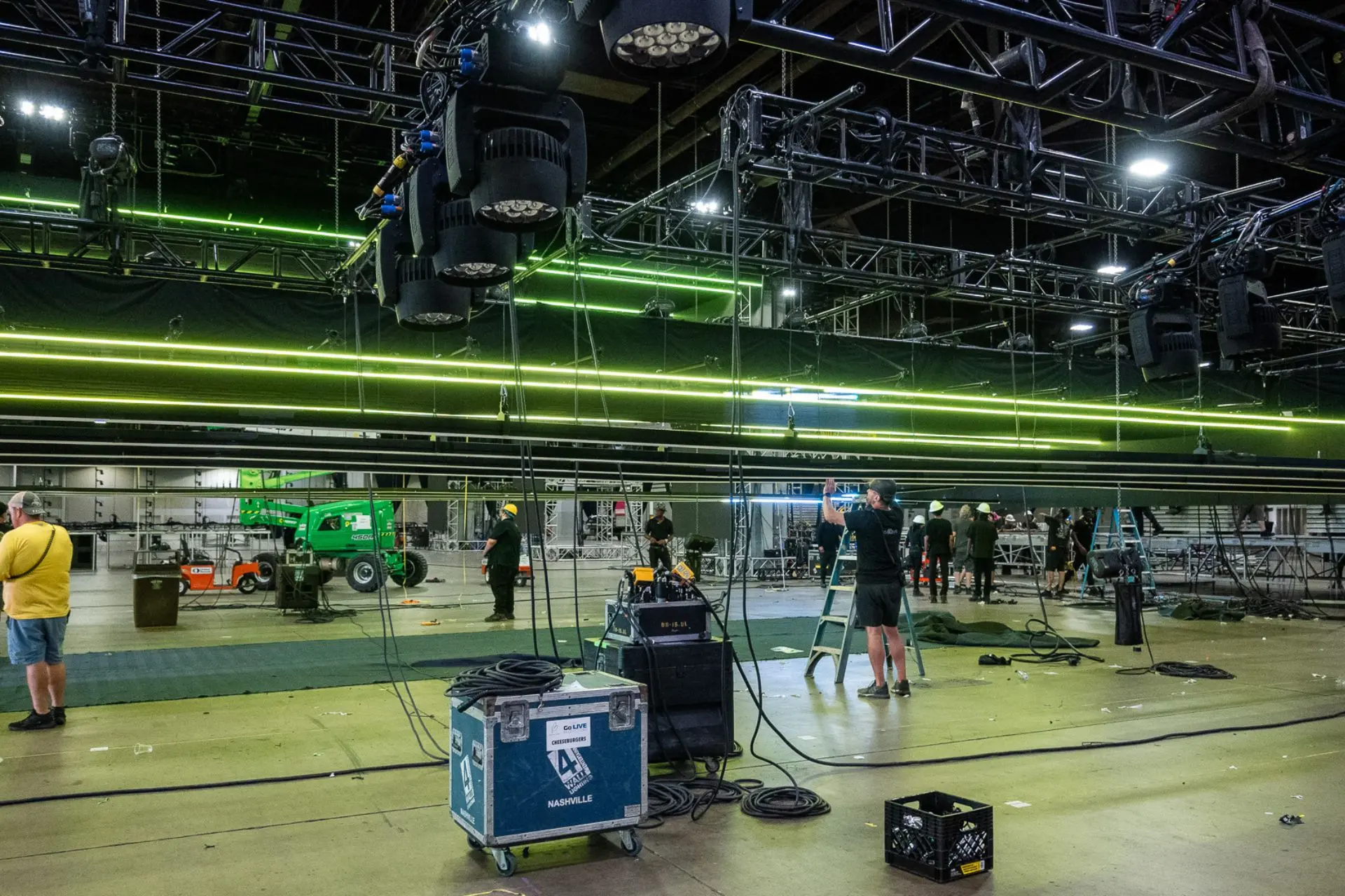 Stage crew setting up lighting and equipment in a large indoor venue with trusses and green neon lights overhead.