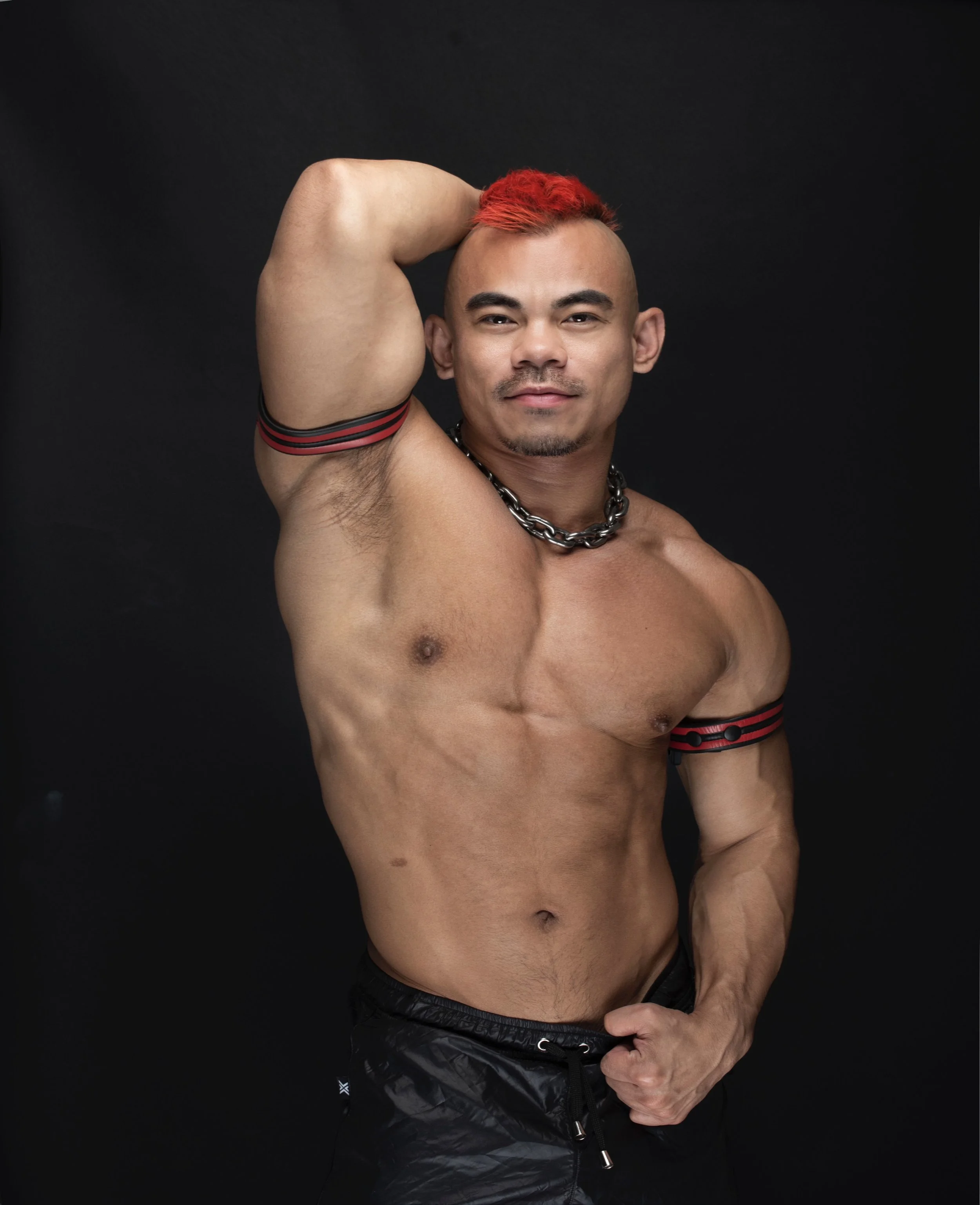 Jason Luna poses with a red mohawk and chain necklace with one arm up showing off his armpit and bicep