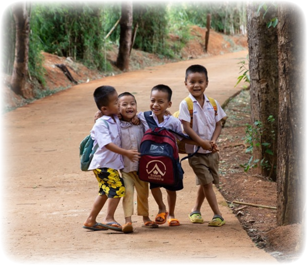 Four young children wearing school uniforms walk together on a rural path in Thailand, smiling and carrying backpacks.