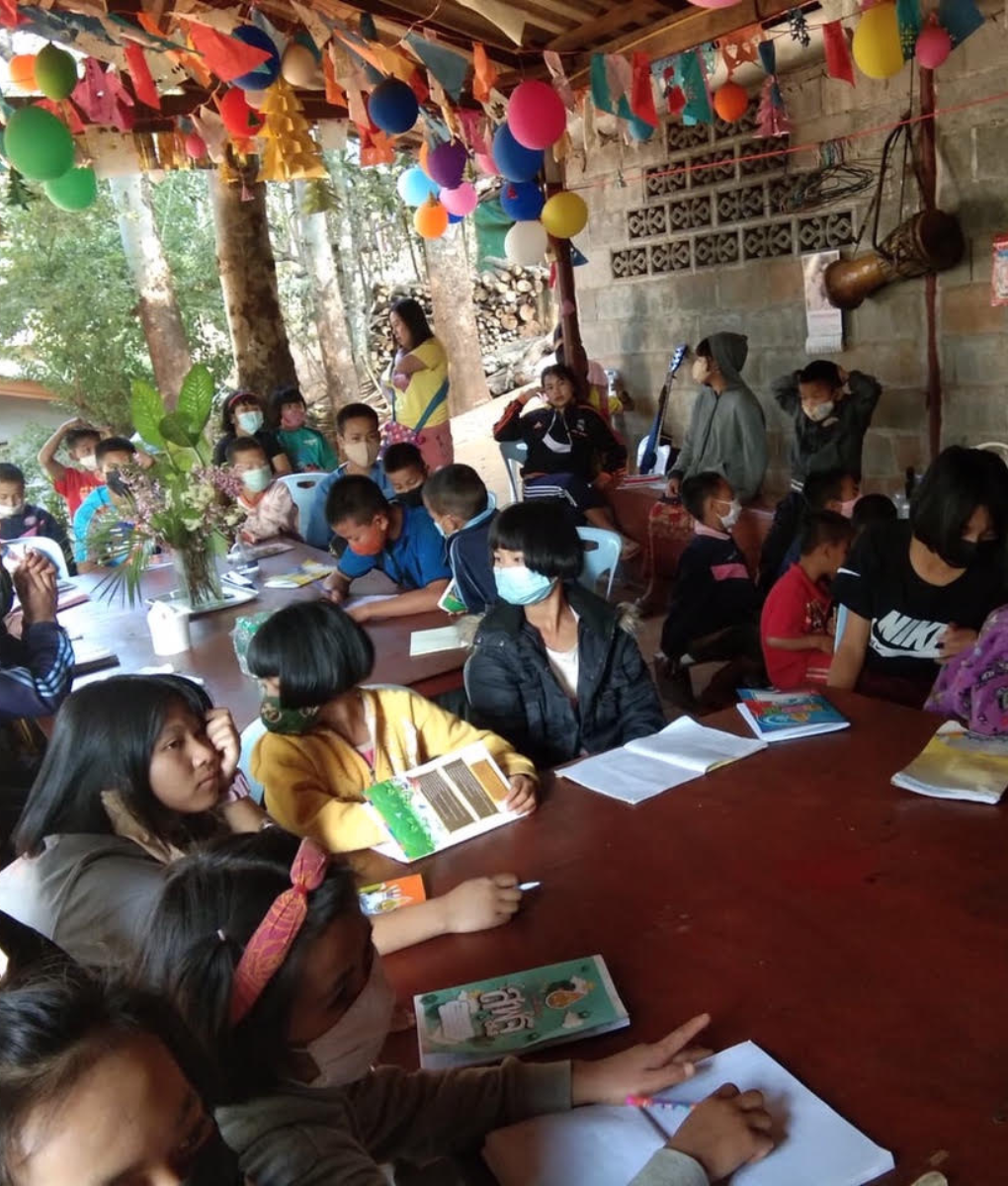 Group of children wearing masks and sitting around tables with books and notebooks in a decorated outdoor classroom.