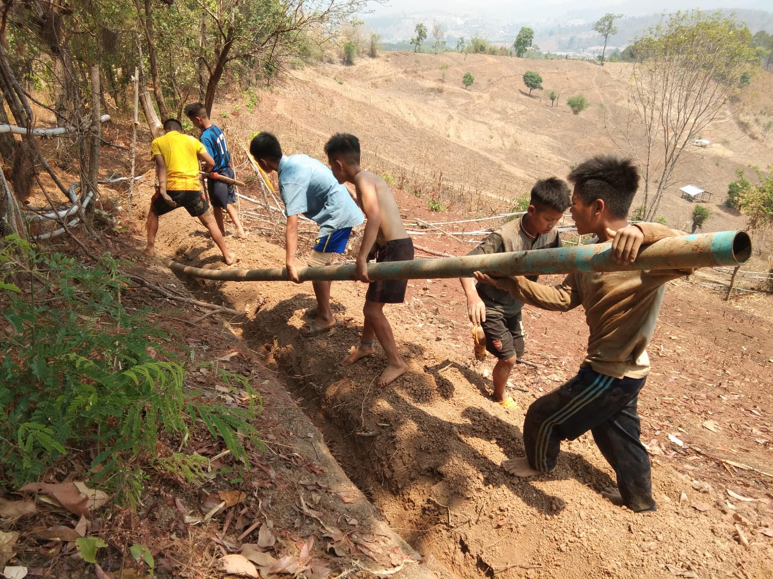 Six boys digging a trench on a dry hillside while carrying a long pipe together.