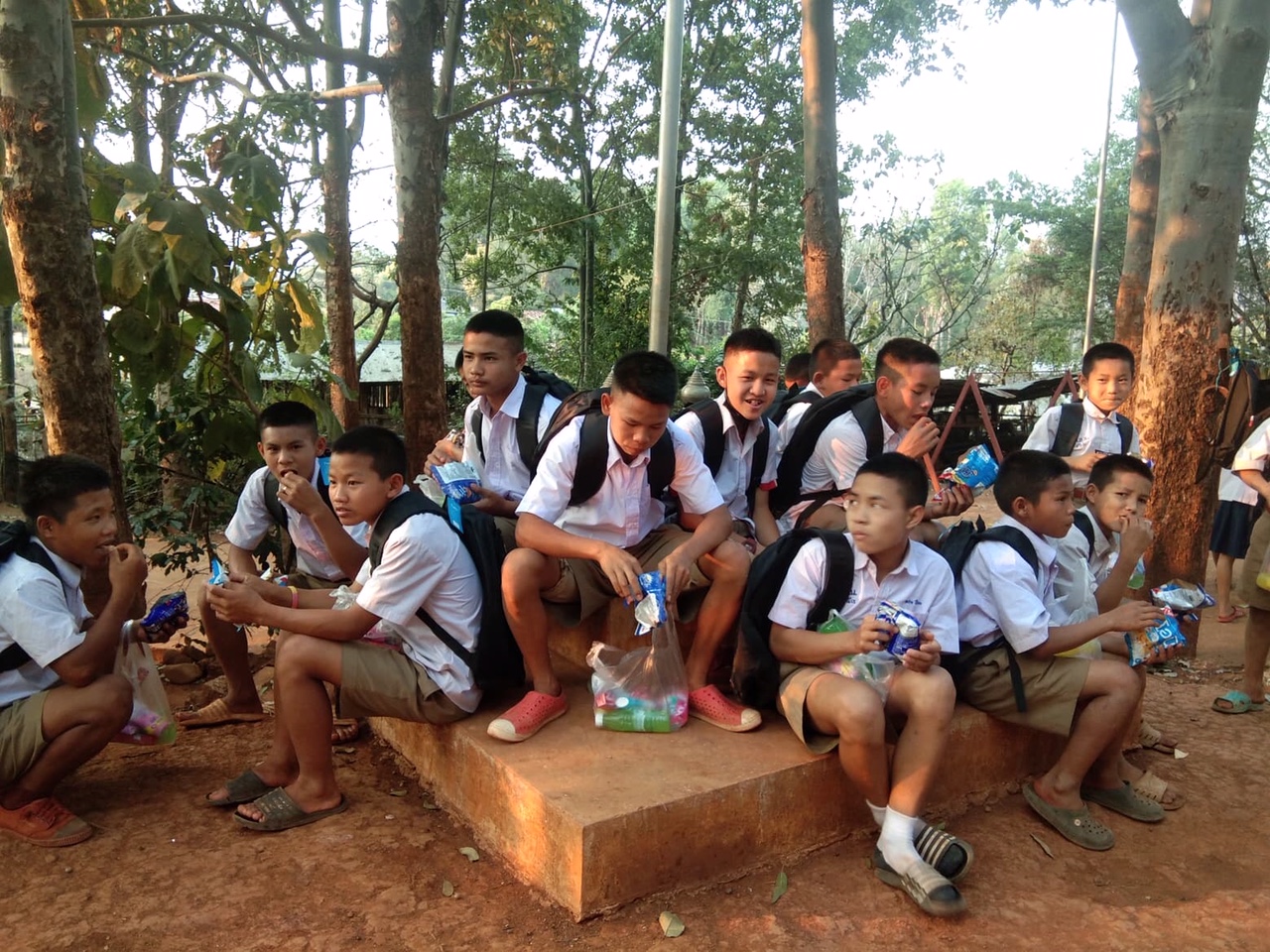 Group of schoolboys in uniform sitting and eating snacks outdoors near trees.