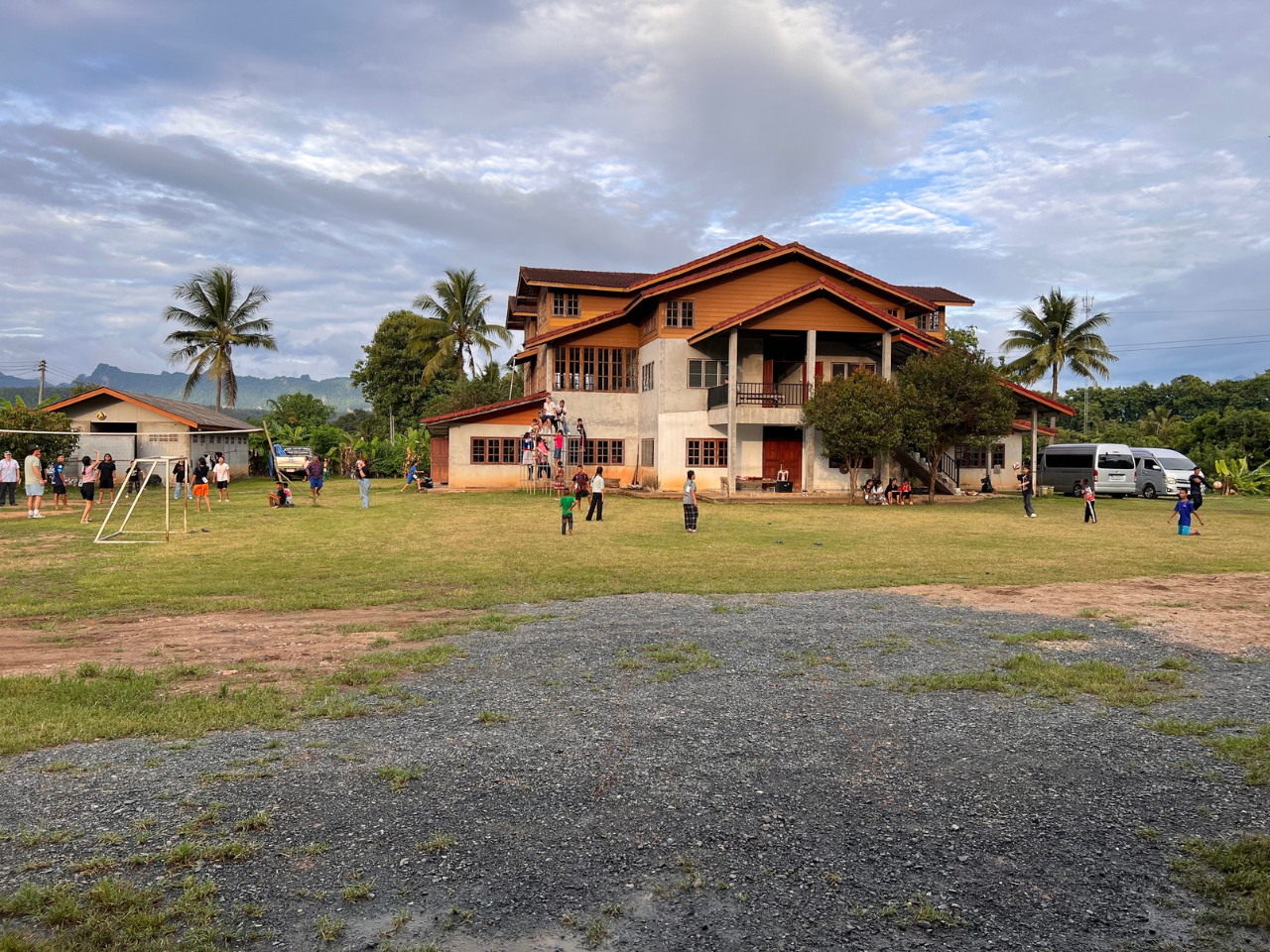 Large group of children and a few adults posing in front of a brick building with a blue roof in a sunny outdoor setting.