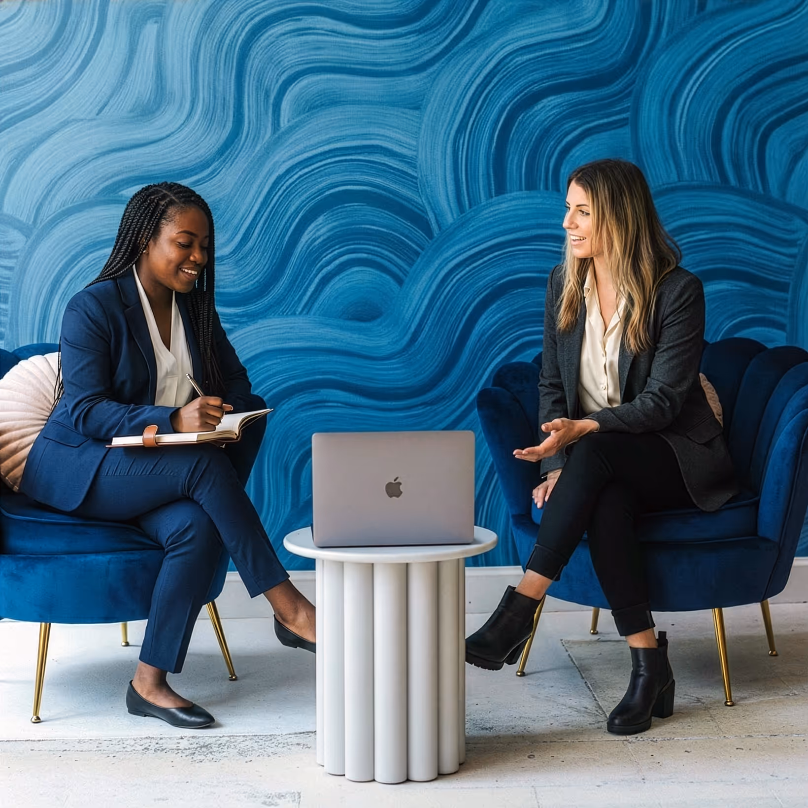 Two women in business attire having a discussion while seated on blue armchairs with a laptop on a small round table between them.