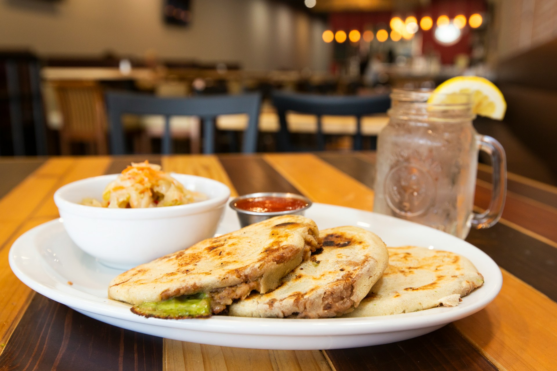 A plate of three pupusas sits on a wooden table, accompanied by a side of slaw and salsa. 