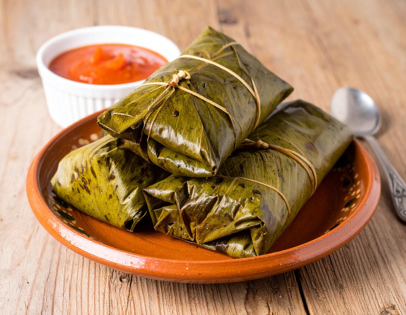 Three tamales wrapped in green banana leaves sit on a clay plate with a small white dish of red sauce in the background. A spoon rests nearby.
