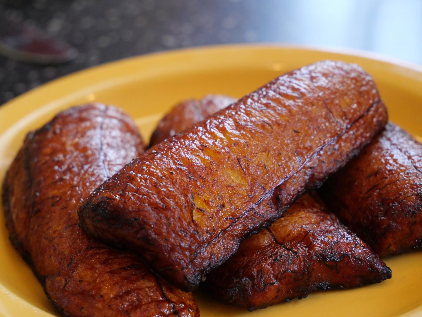 Close-up of golden-brown fried plantains on a yellow plate. The plantains appear caramelized and crispy.