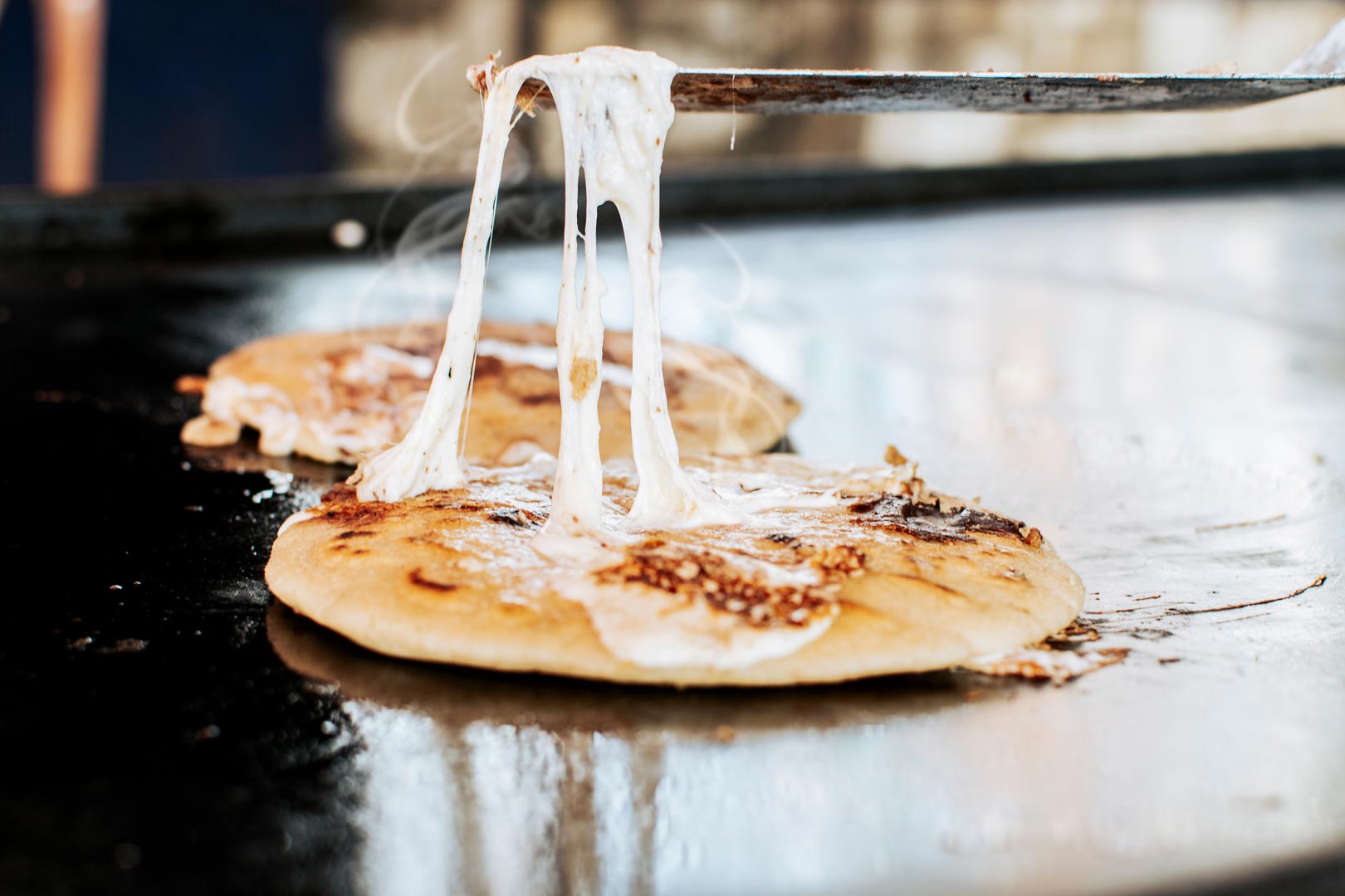 A close-up of a melted cheese pupusa on a griddle, being lifted by a spatula. 