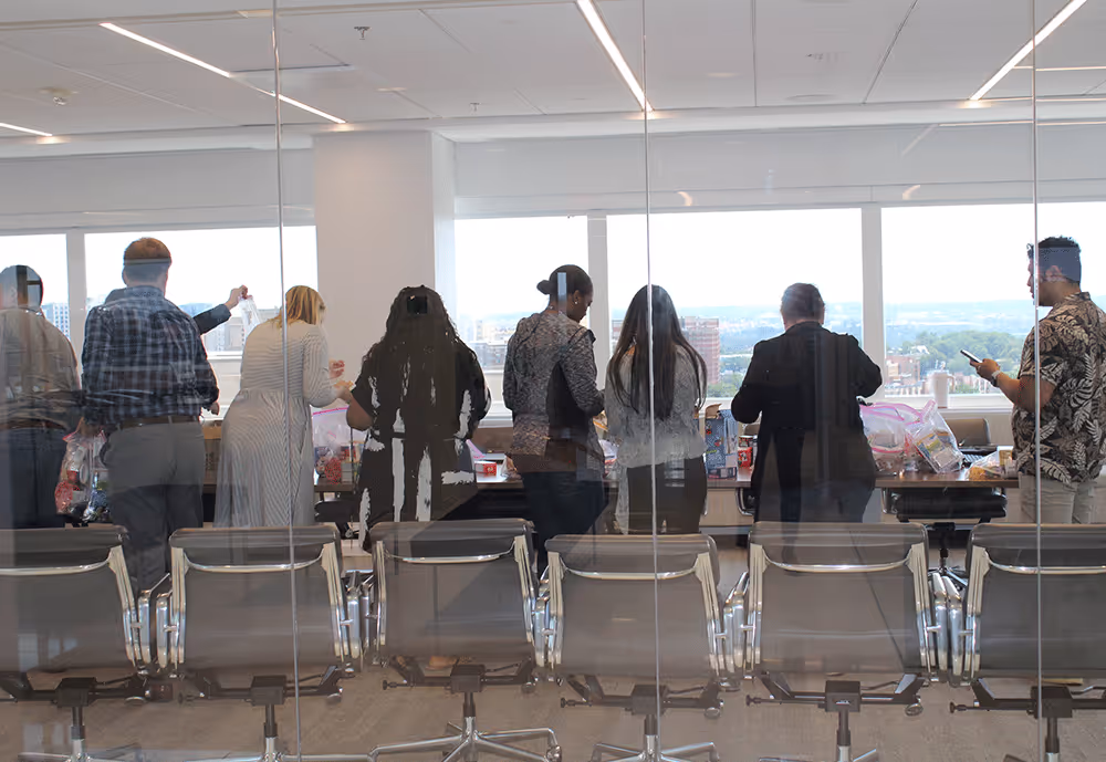 Group of people standing and working together at a table in a modern office with large windows overlooking a city.