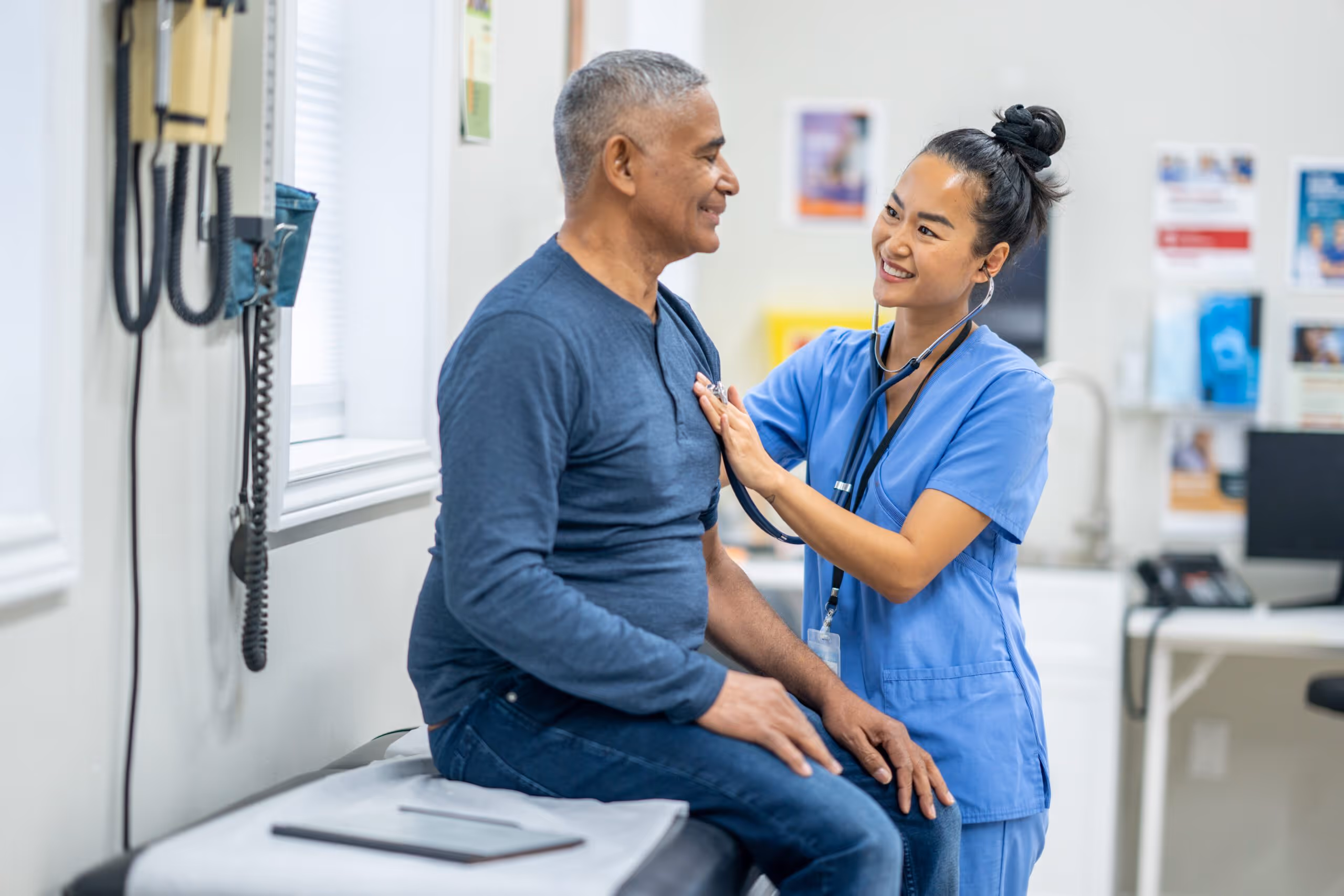 Healthcare professional uses a stethoscope to examine a smiling male patient sitting on an exam table.