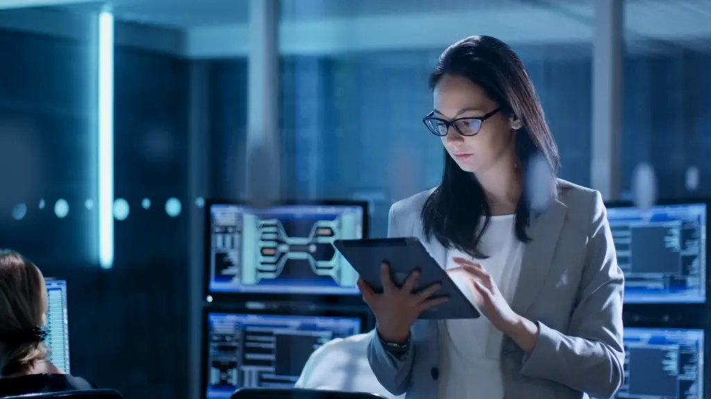 Woman in glasses using a digital tablet in a dimly lit room with multiple computer screens displaying data and diagrams.