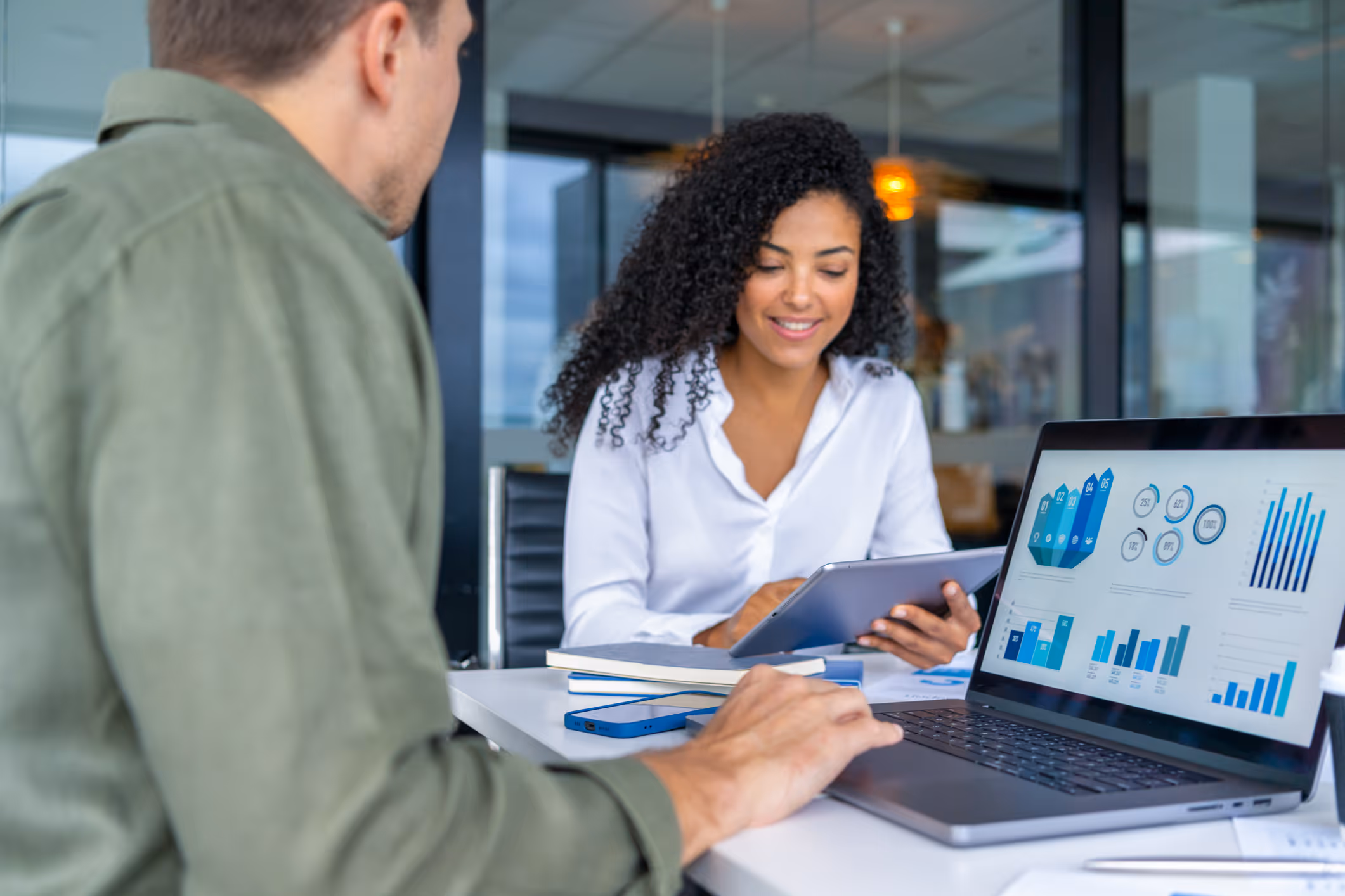 Two professionals in an office discussing data on a tablet and a laptop screen displaying charts and graphs.
