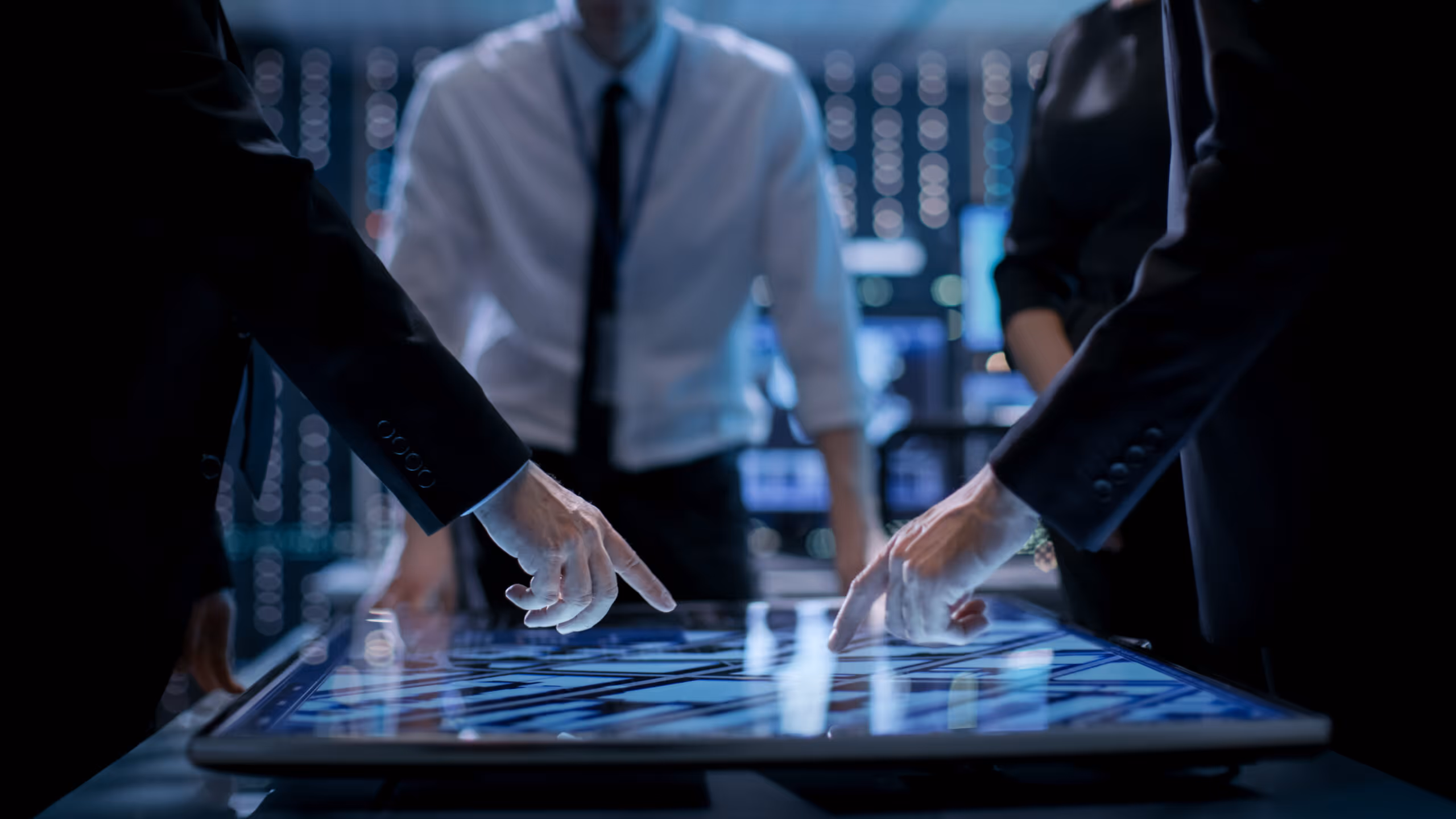Business professionals pointing at data on a large interactive touchscreen table in a dimly lit office.