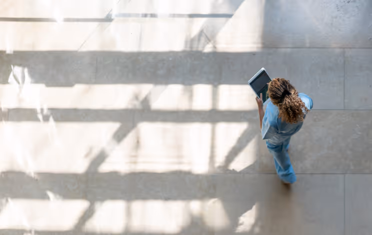 Top view of a medical professional in blue scrubs walking and holding a laptop in a sunlit hallway.