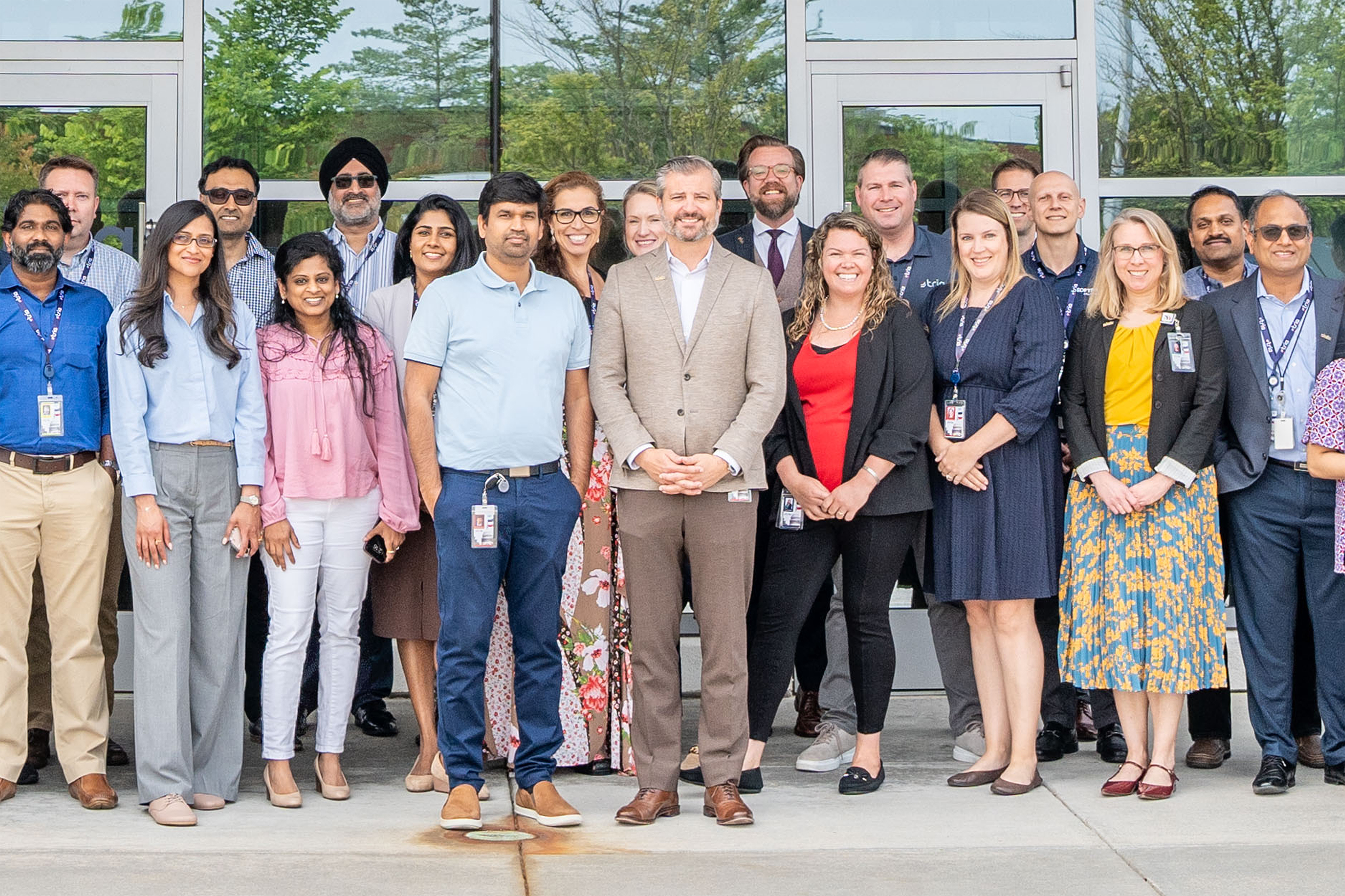 Group of diverse professionals standing in front of a modern office building with glass windows, flanked by two signs for Tria, highlighting their federal health ecosystem solutions.