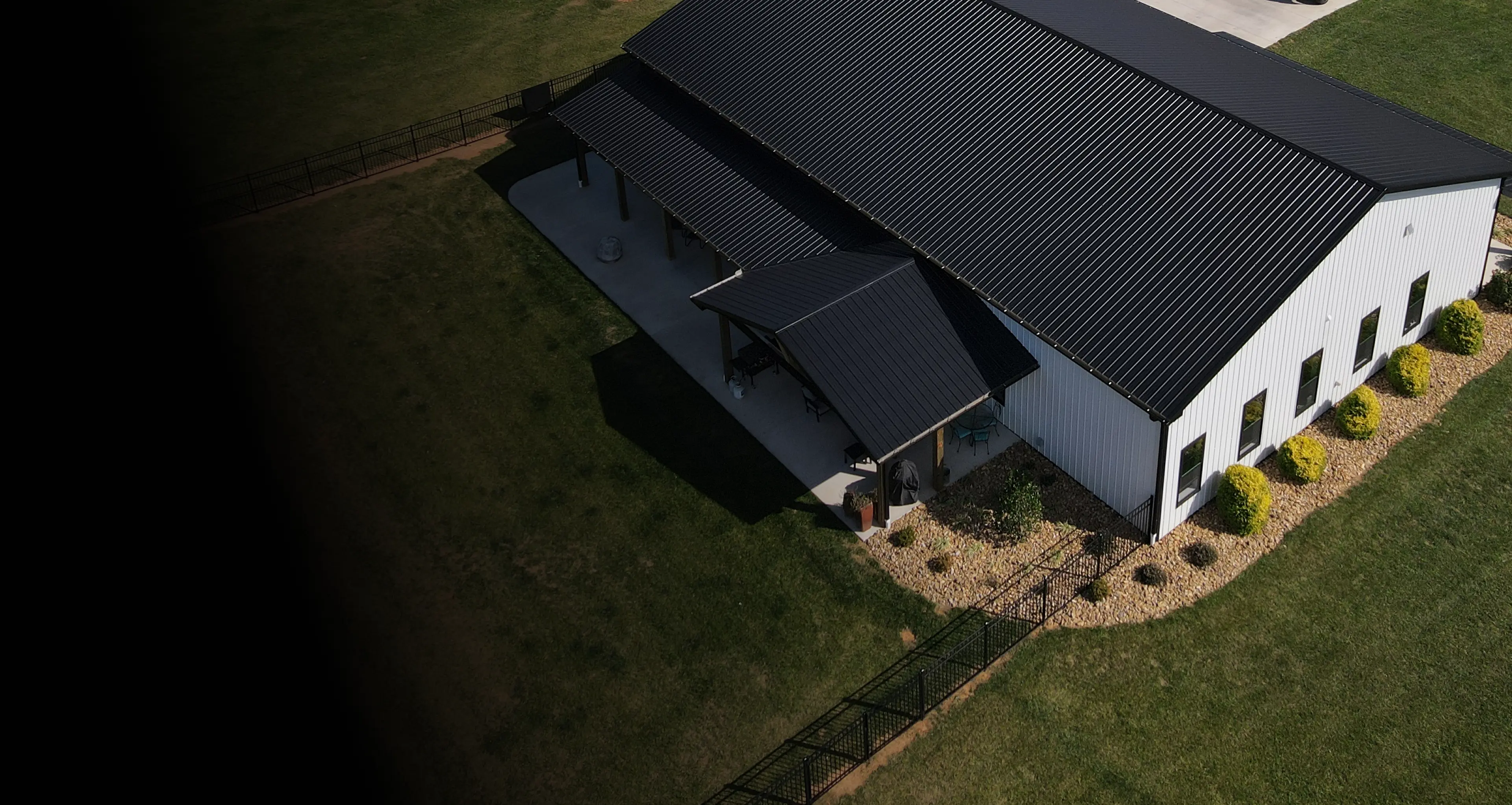 Aerial view of a modern white barn with a black metal roof, surrounded by grass, landscaping, and a paved driveway.