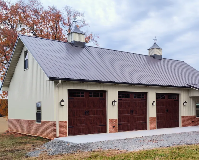 Large beige barn with brick base, gray metal roof, and three dark wooden garage doors under a cloudy sky.