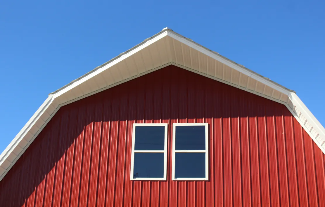 Red barn wall with white-trimmed double windows under a white roof against a clear blue sky.