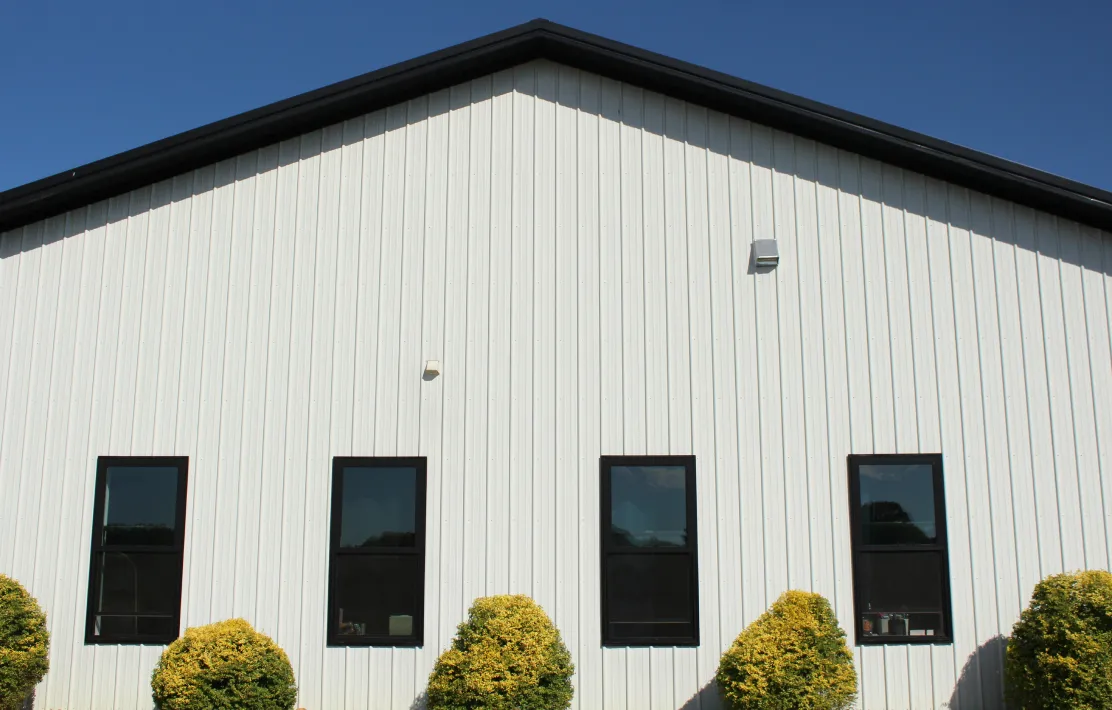 White metal building facade with four vertically aligned black-framed windows and trimmed bushes in front.