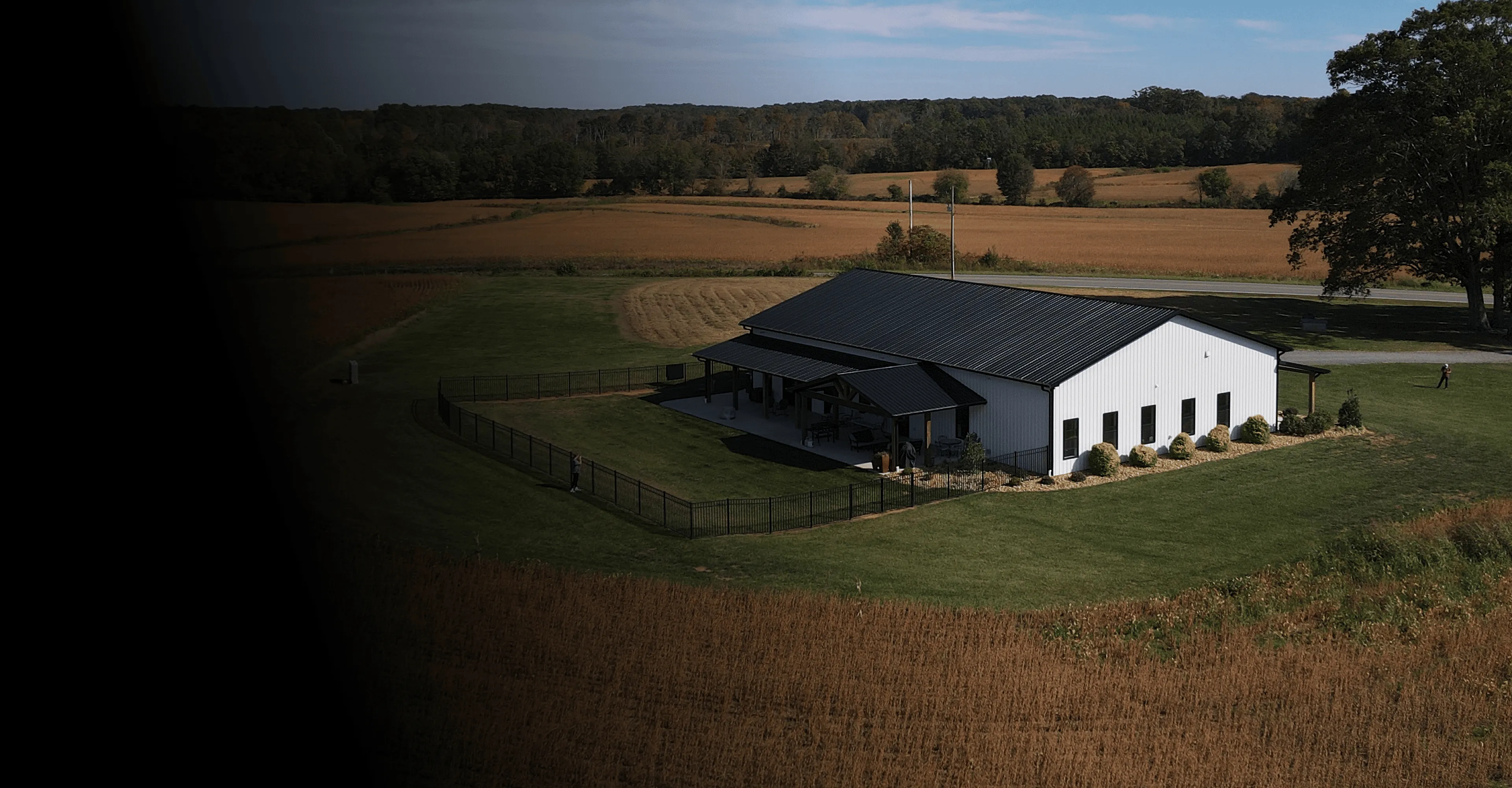 Aerial view of a white building with a black roof surrounded by a fenced green lawn, with farmland and trees in the background.