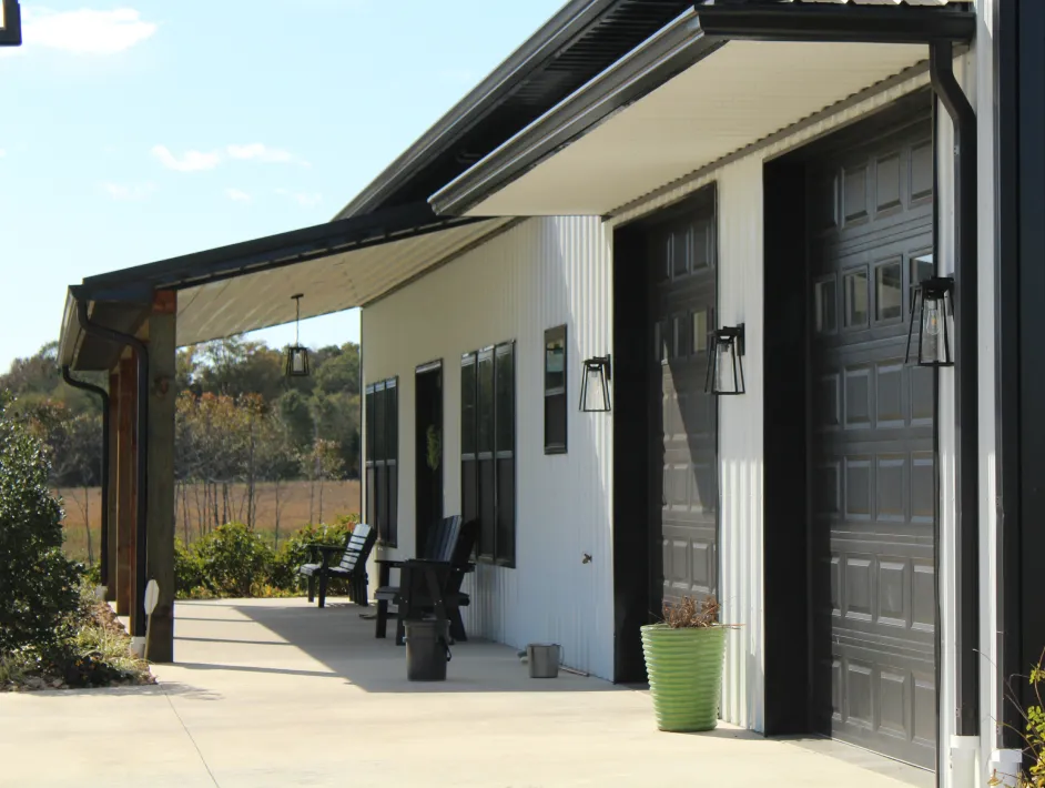 Side view of a white building with black garage doors, black window frames, outdoor wall lanterns, and a concrete patio with black chairs and green plants.