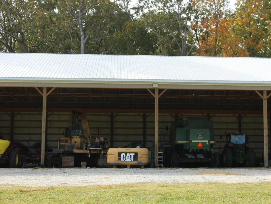 Open-sided metal storage shed housing various farm machinery including a CAT excavator and green John Deere equipment.