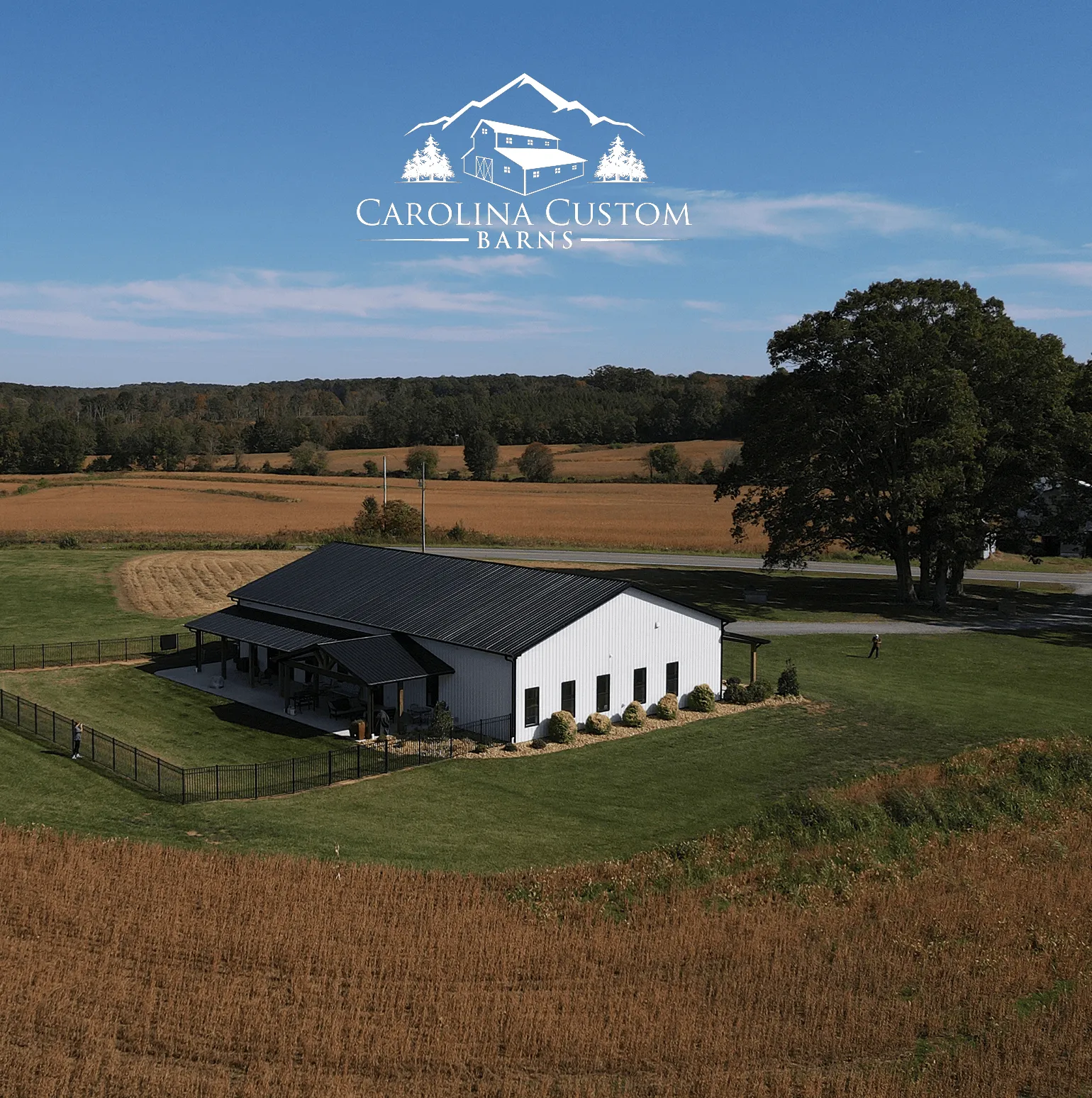 A modern white barn with a black roof surrounded by green grass and brown fields under a clear blue sky, with the Carolina Custom Barns logo above.