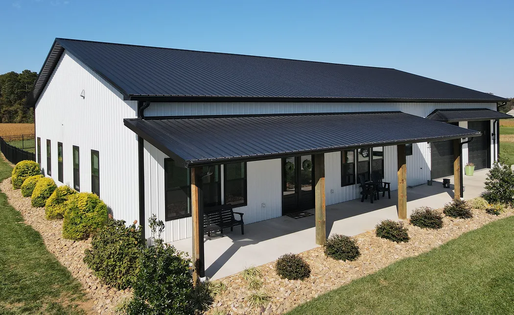 Modern white barn with a black metal roof, front porch with wooden posts, and landscaped shrubs and rocks along the sides.