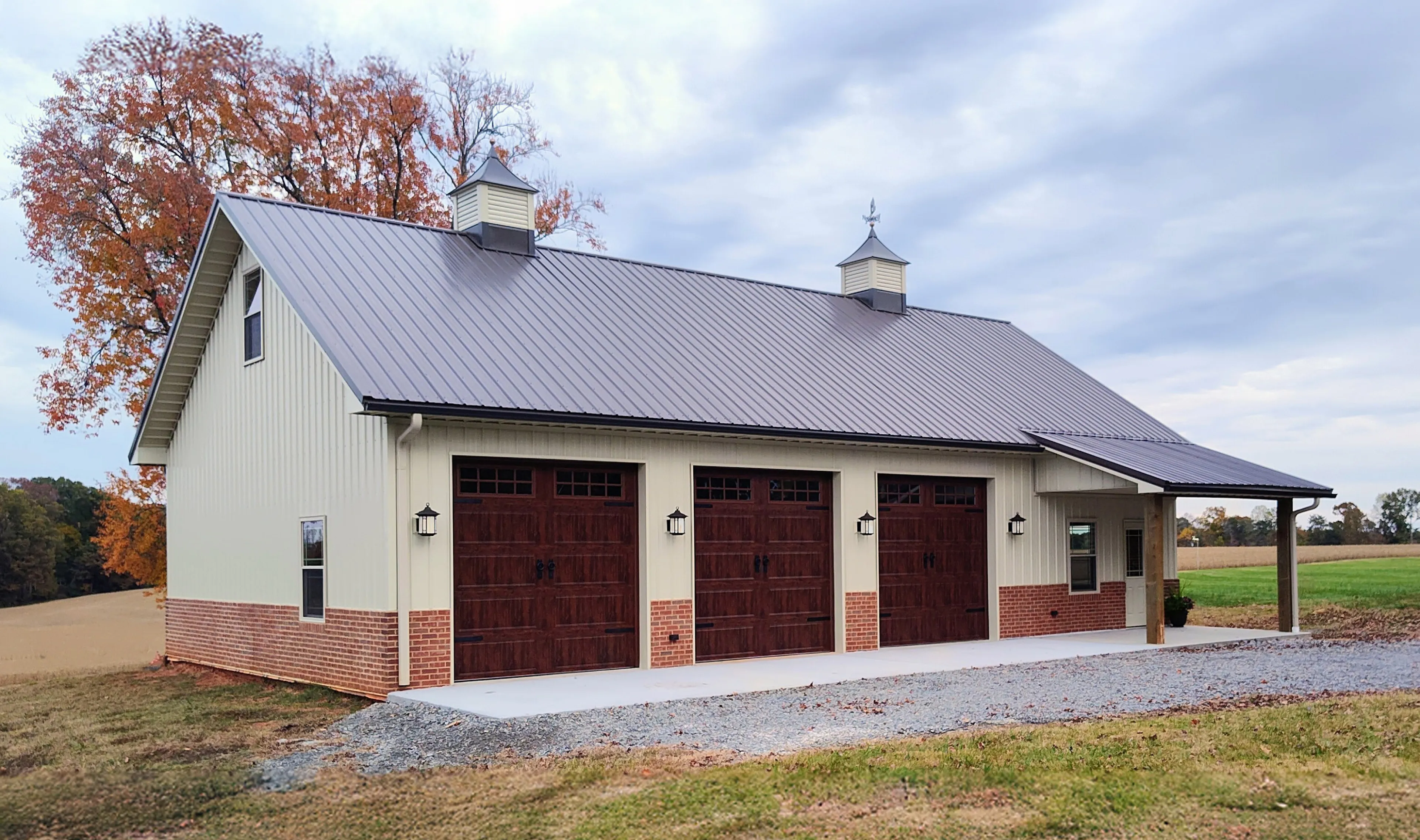 Large detached garage with three wooden doors, metal roof, and small cupolas, surrounded by grass and trees.