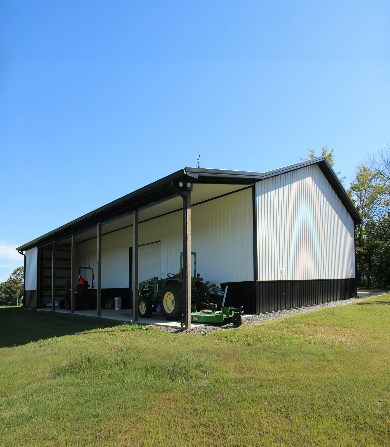 Large metal agricultural shed with open side housing a tractor and equipment on grassy land.