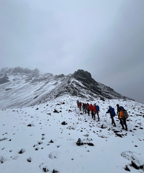 Group of hikers trekking up a snow-covered mountain slope under a cloudy sky.