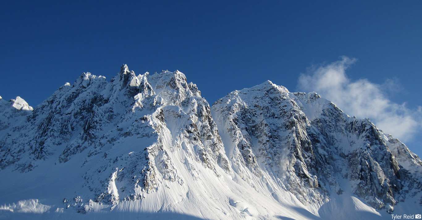 Snow-covered rugged mountain peaks under a clear deep blue sky with wisps of clouds.