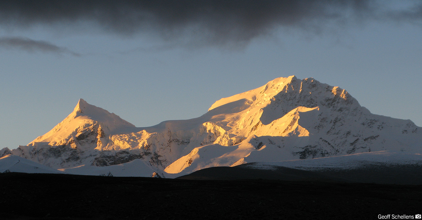 Sunlit snow-covered mountain peaks under a dark, cloudy sky at dusk.