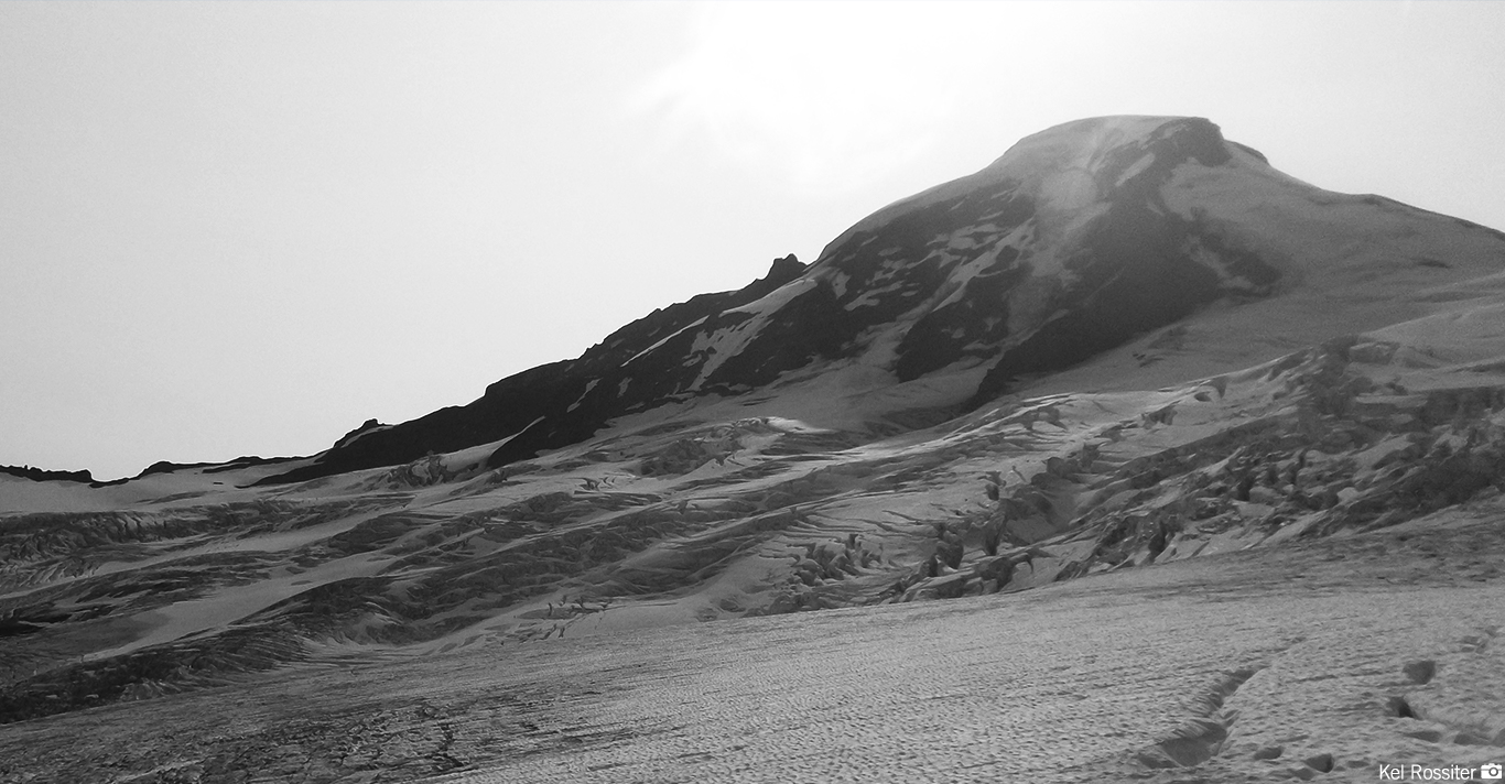 Snow-covered mountain peak with rugged ridges under a bright, overcast sky in black and white.