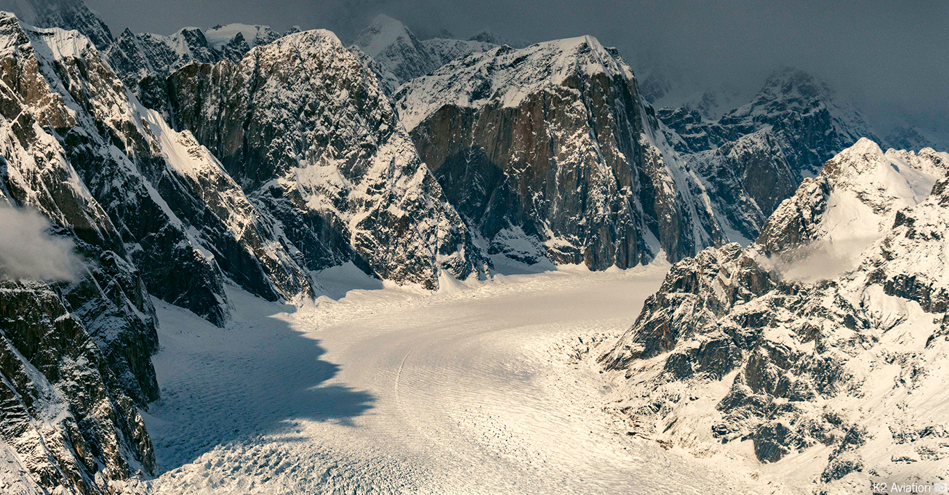 Snow-covered rugged mountain peaks surrounding a wide glacier under a cloudy sky.