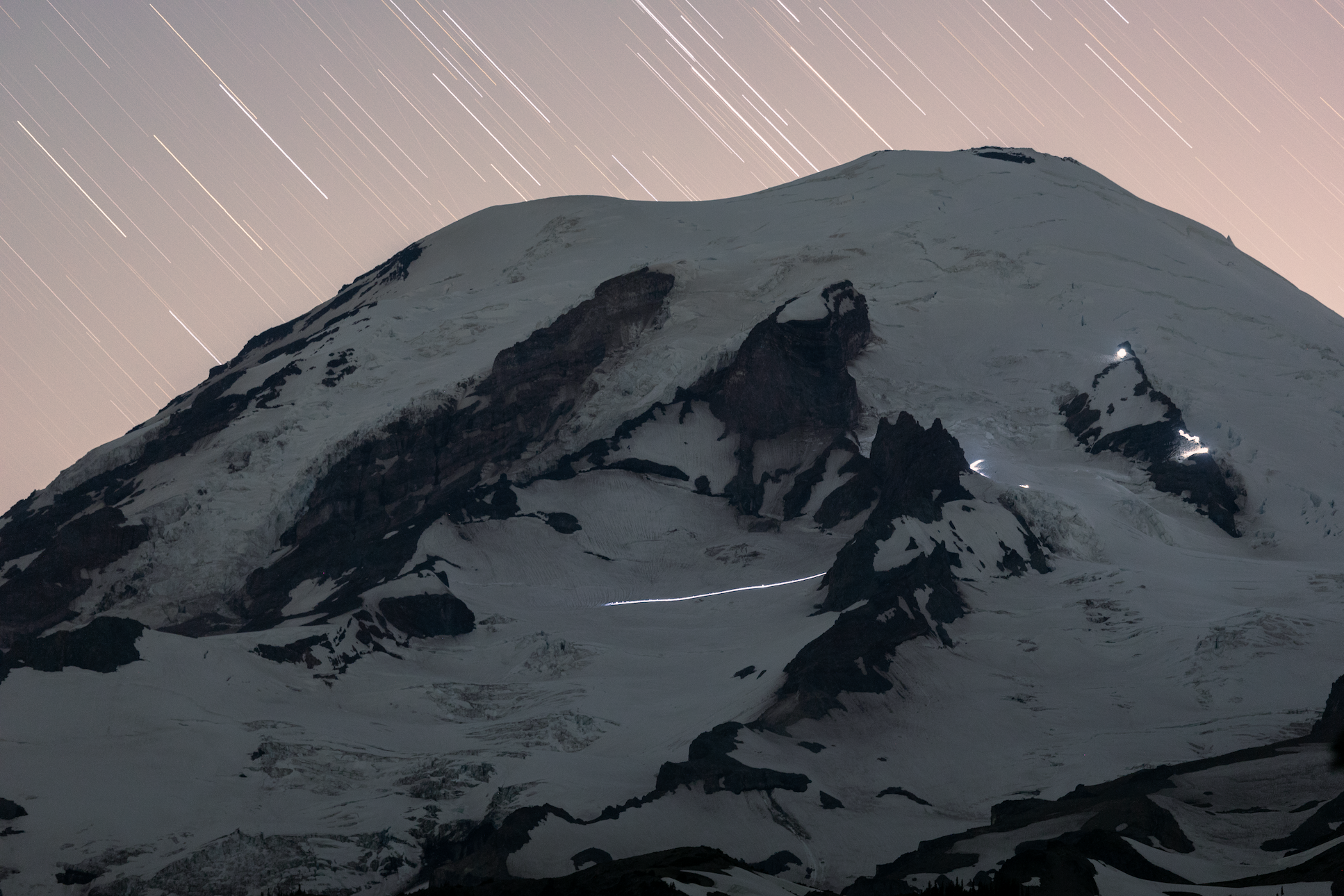 Snow-covered mountain under night sky with star trails and climbers' headlamps lighting the slope.