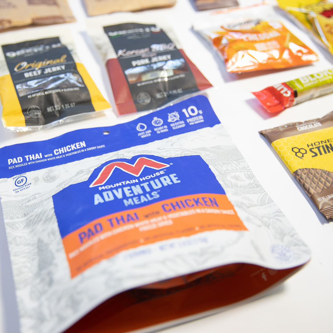 Mountain House Adventure Meals Pad Thai with chicken package among various snack packets on white surface.
