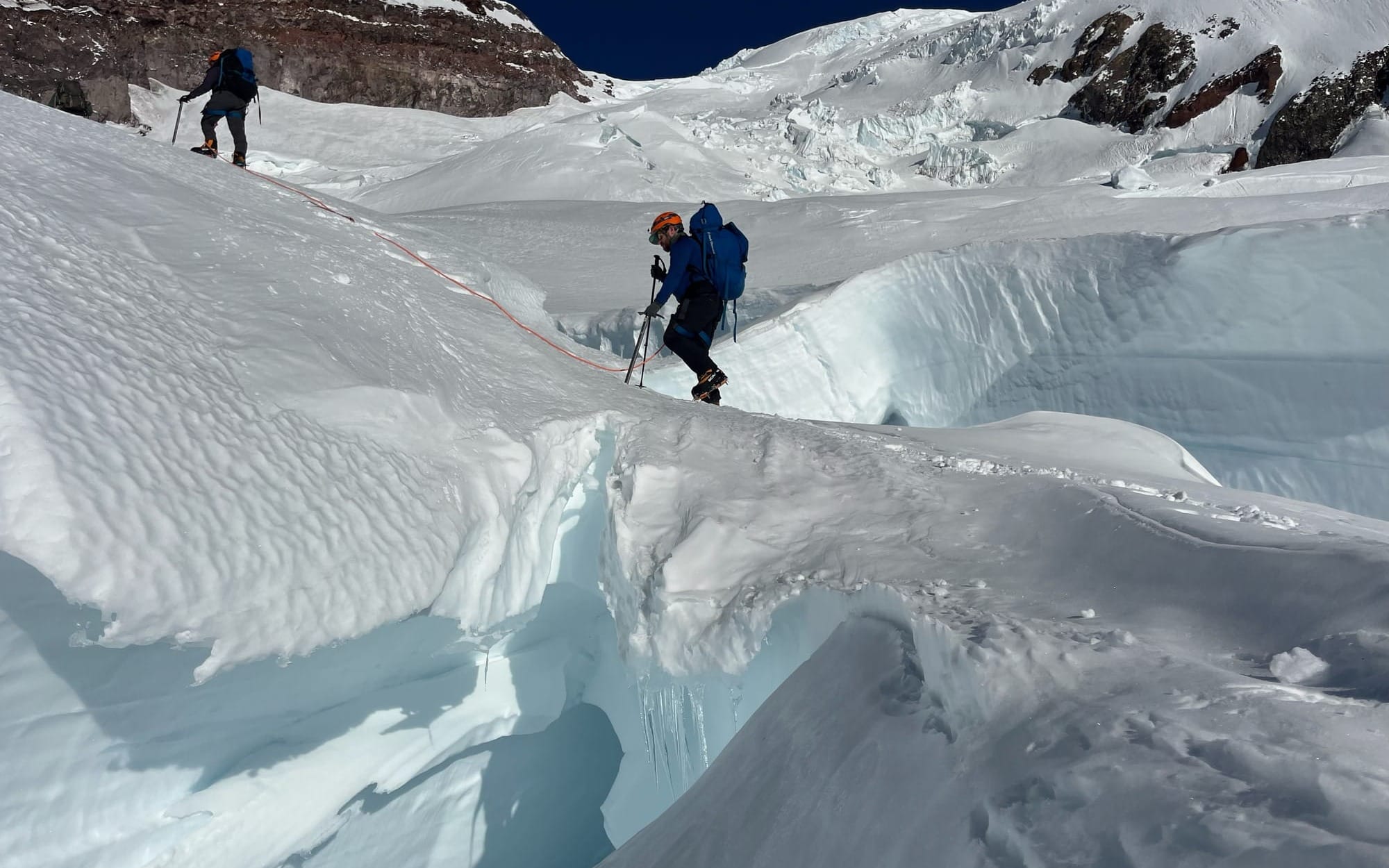 Two climbers in blue gear crossing a narrow snow bridge over a deep crevasse on a glacier.